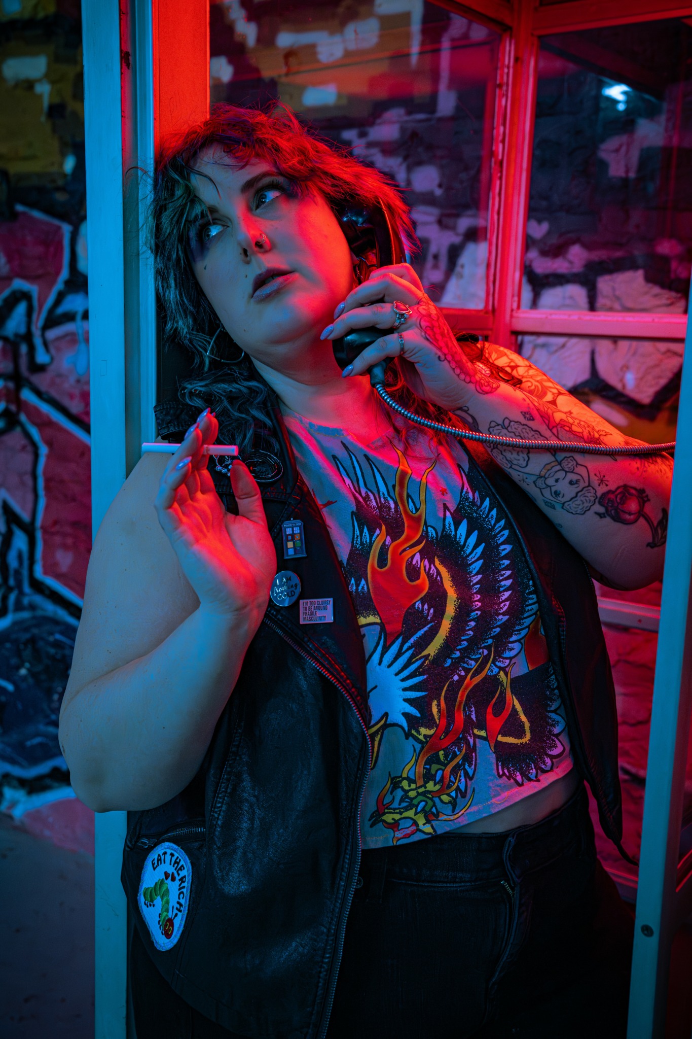 Young woman with curly hair and tattoos, wearing a graphic shirt and leather vest, in a colorful, graffiti-covered setting.