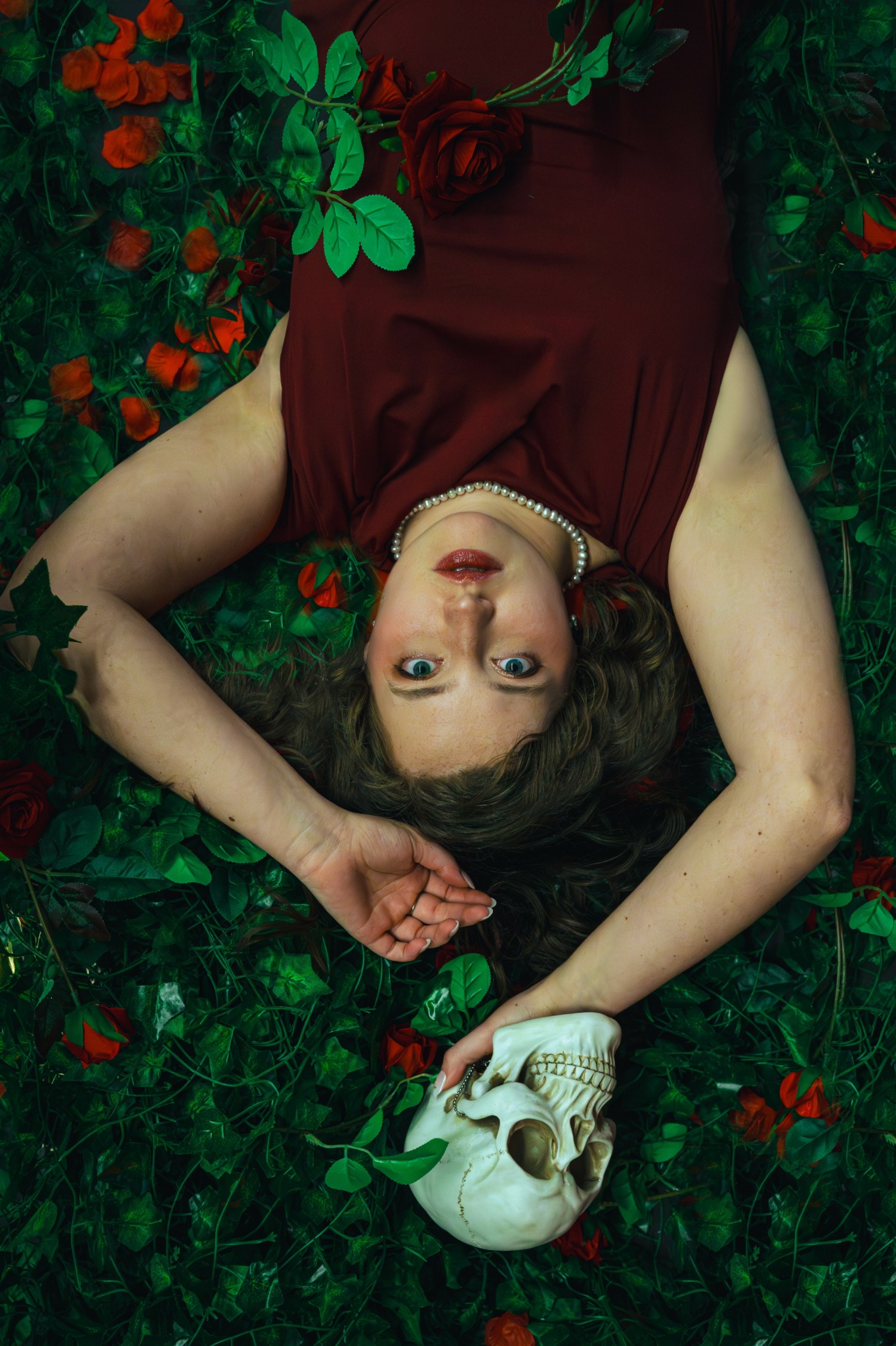 Woman lying on ground surrounded by greenery and red flowers, holding a skull in her hand, looking up.