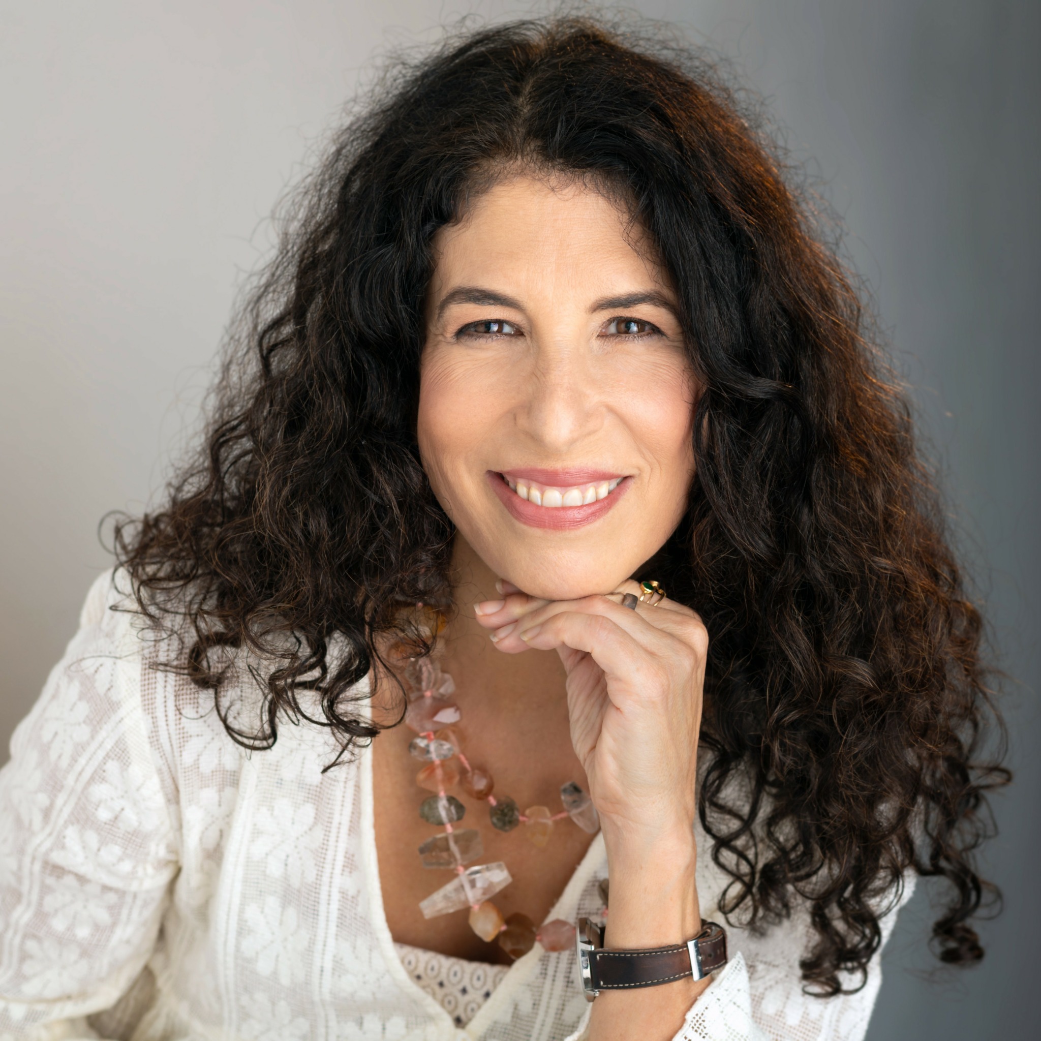 Woman with curly dark hair smiling, resting chin on hand, wearing a white top and necklace.