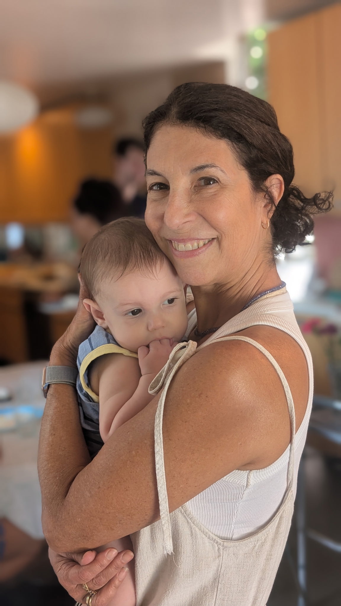 Woman holding a baby indoors, smiling, with blurred background of people and warm lighting.