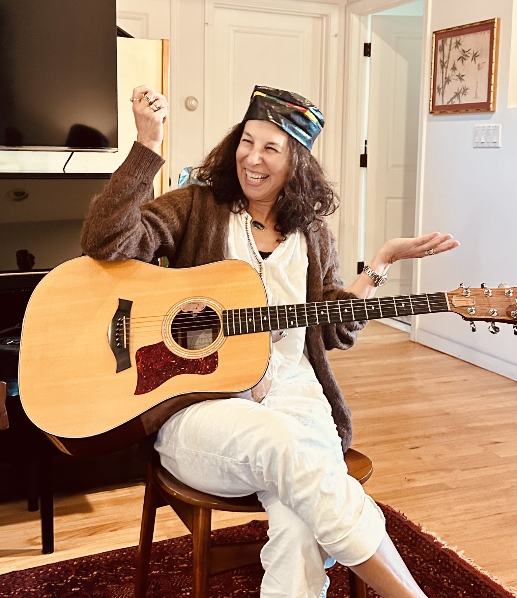 Woman sitting on a chair holding an acoustic guitar, smiling, wearing a colorful hat and casual clothing in a living room.