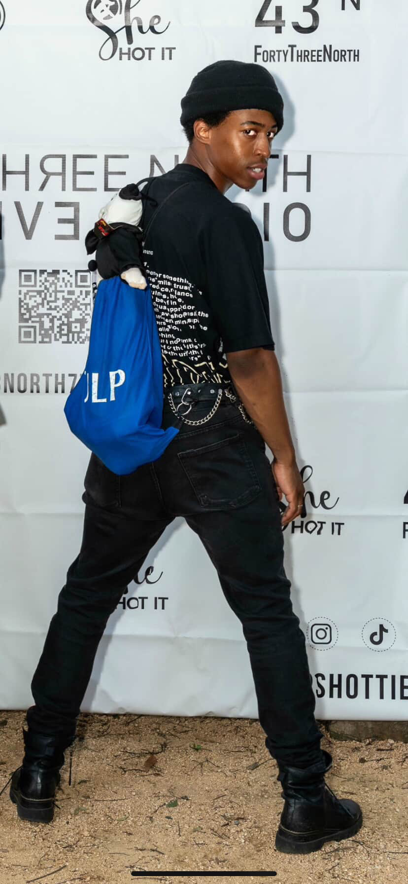 Young person with black beanie and black outfit, carrying a blue bag, standing against a white backdrop.