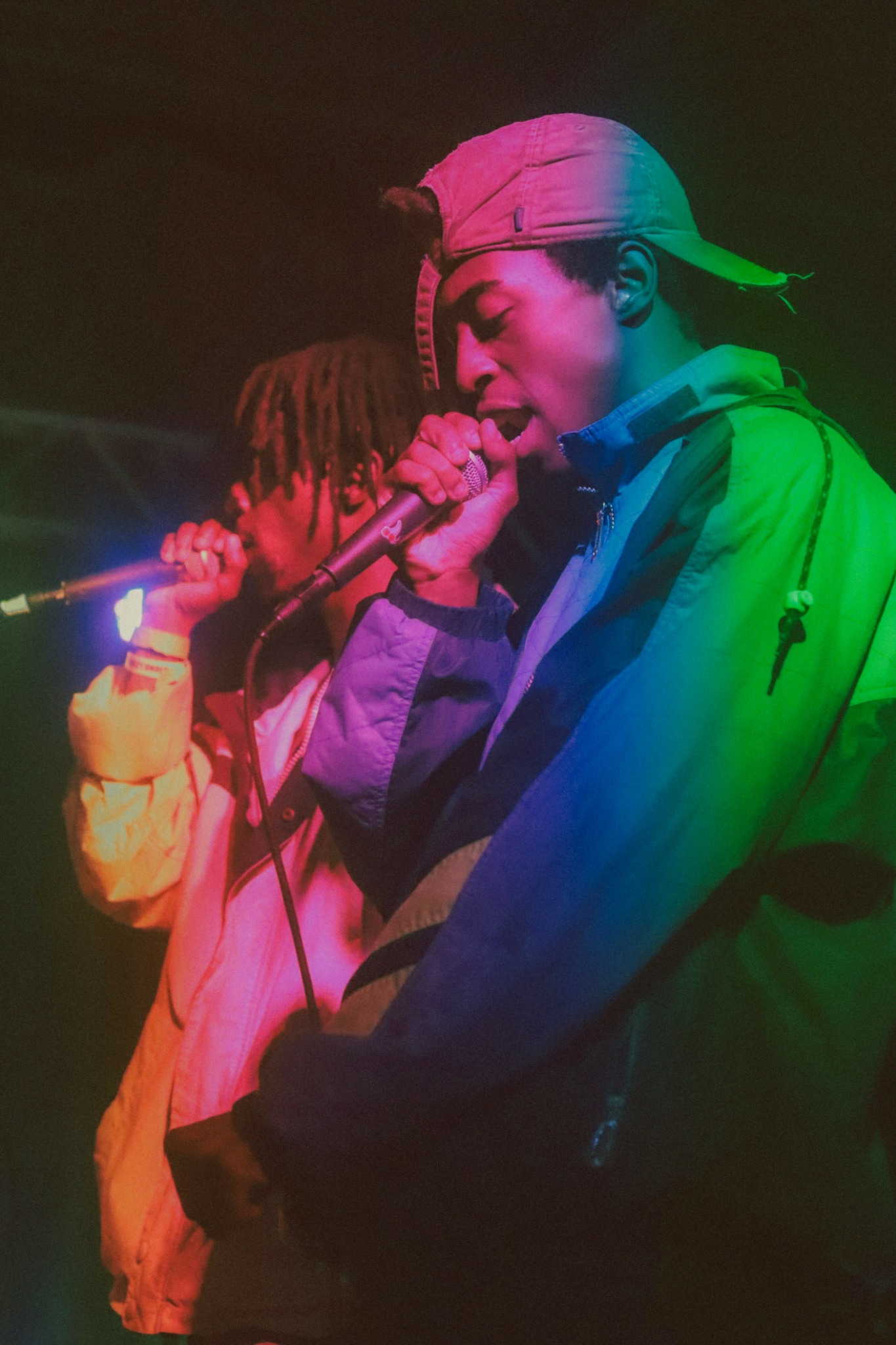 Two people singing into microphones on stage, one wearing a cap and jacket, colorful lighting effects.