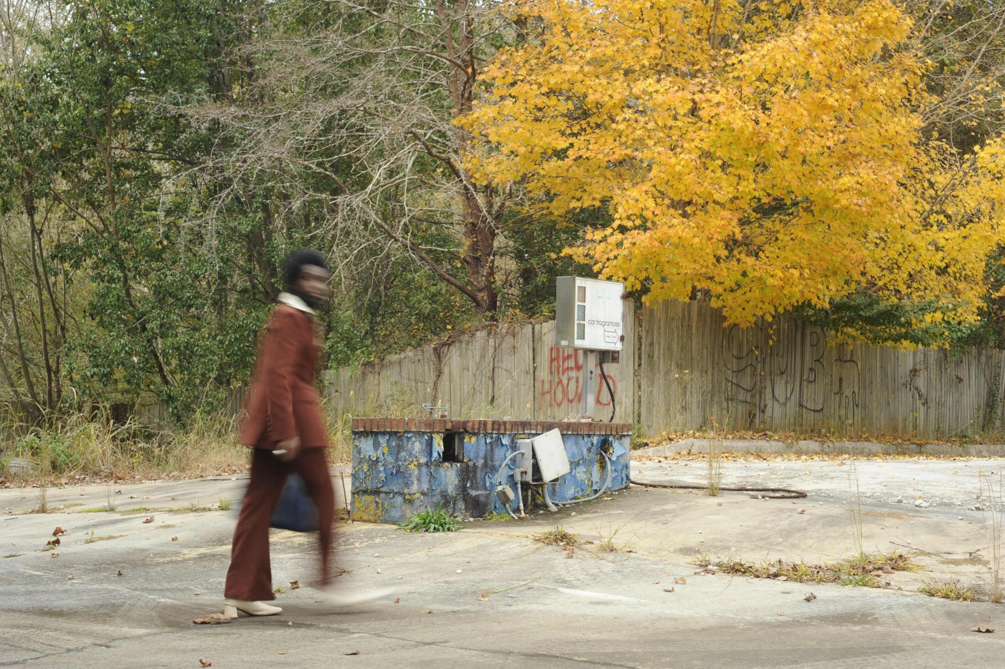 Person walking past a blue, weathered water fountain with autumn trees and a wooden fence in the background.
