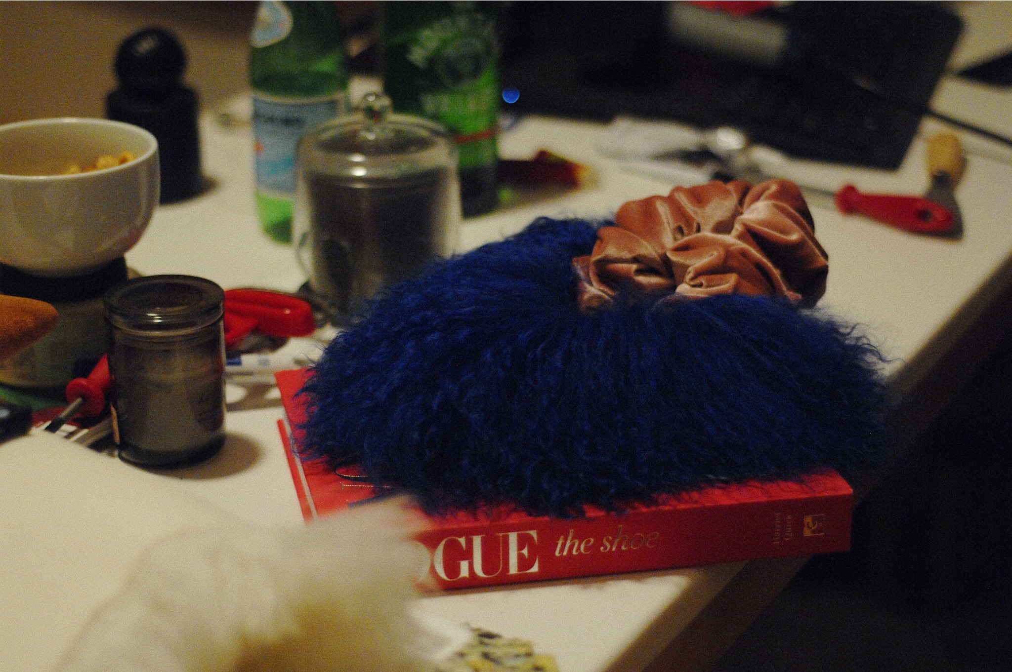 Blue fuzzy object with a brown scrunchie on top, placed on a red book on a cluttered table.