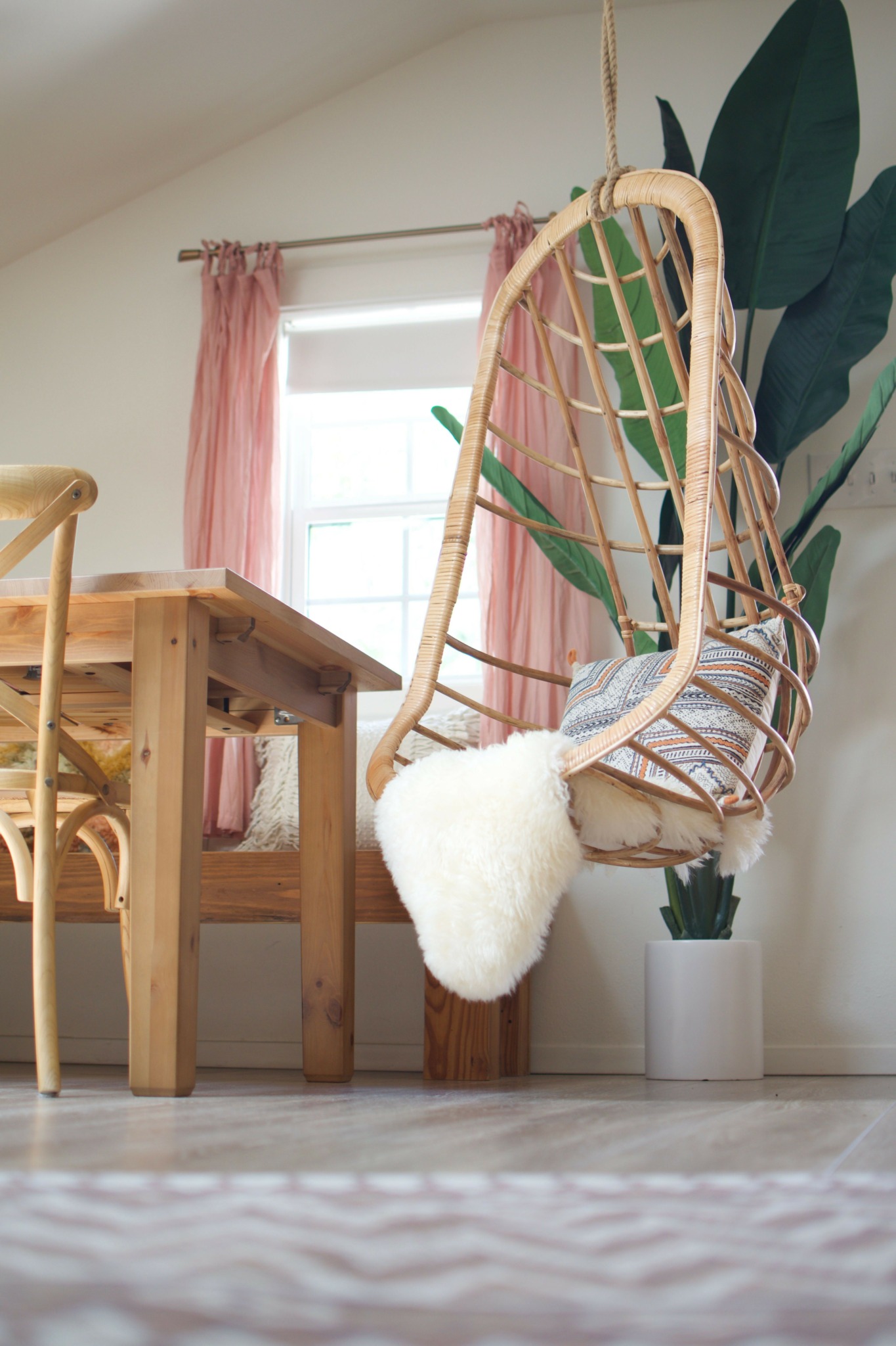 Wicker chair with green cushion and large plant in a room with pink curtains and a window.