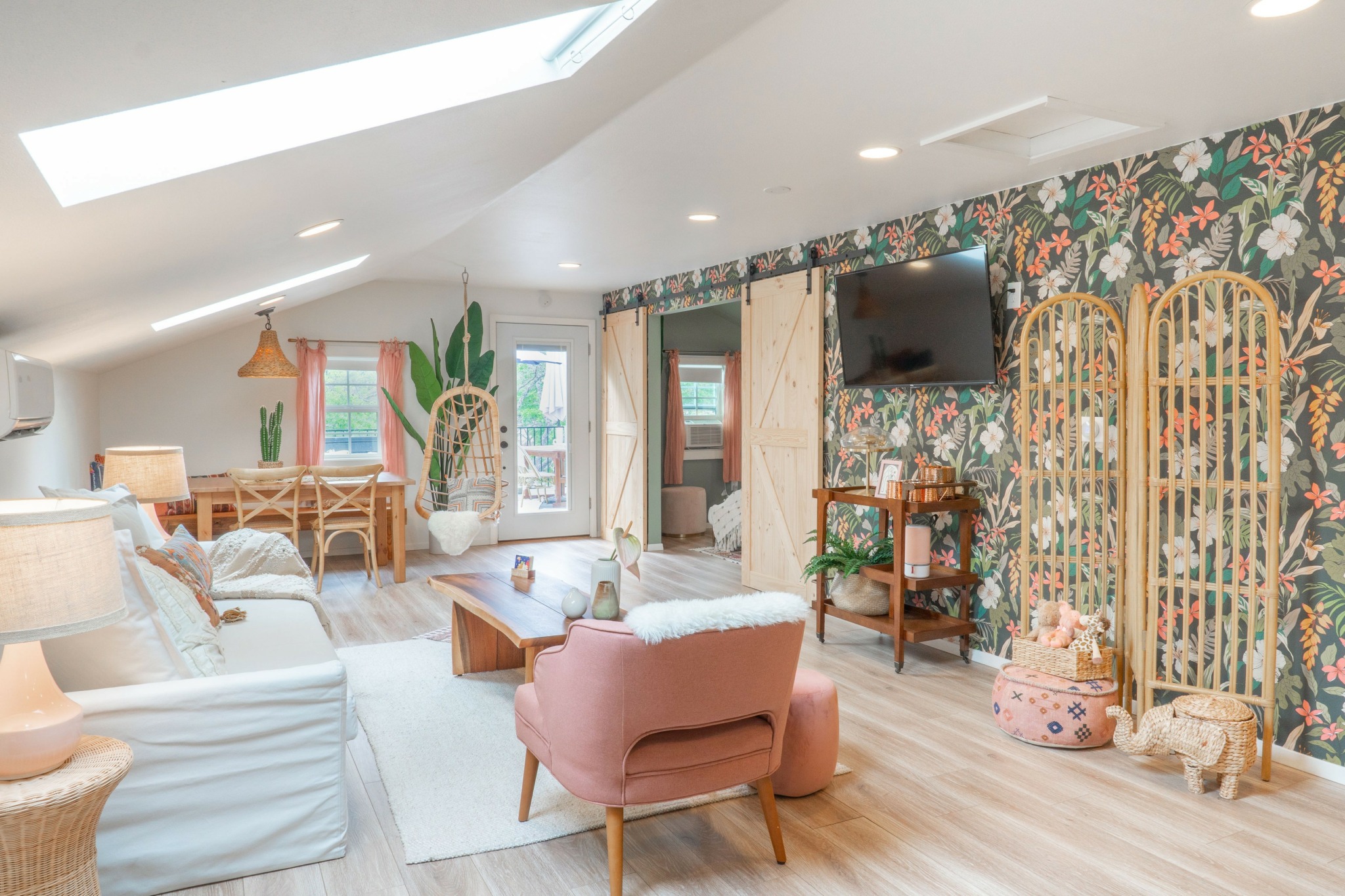 Living room with pink armchair, white sofa, floral wallpaper, TV, and wooden furniture, illuminated by skylight and natural light.