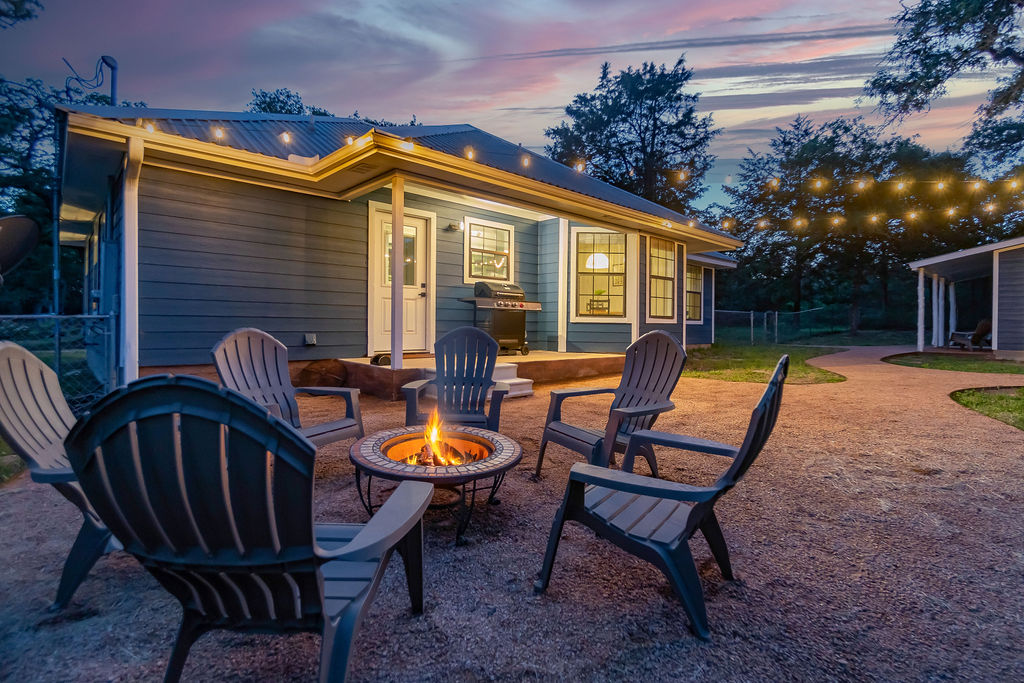 Outdoor backyard with a house, fire pit, and chairs, illuminated by string lights at dusk.