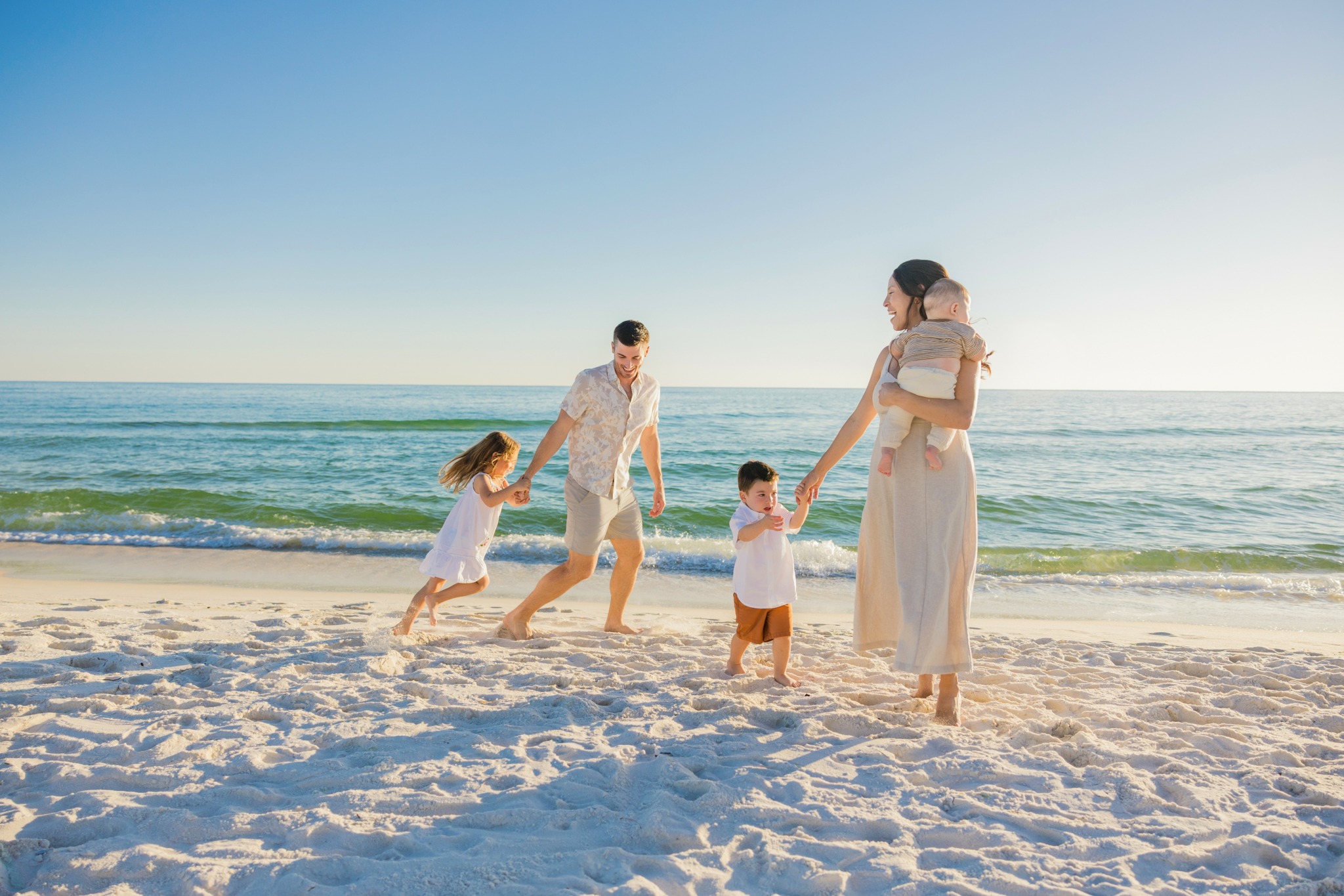 Family of four on beach, children playing, adults holding hands, ocean and clear sky in background.