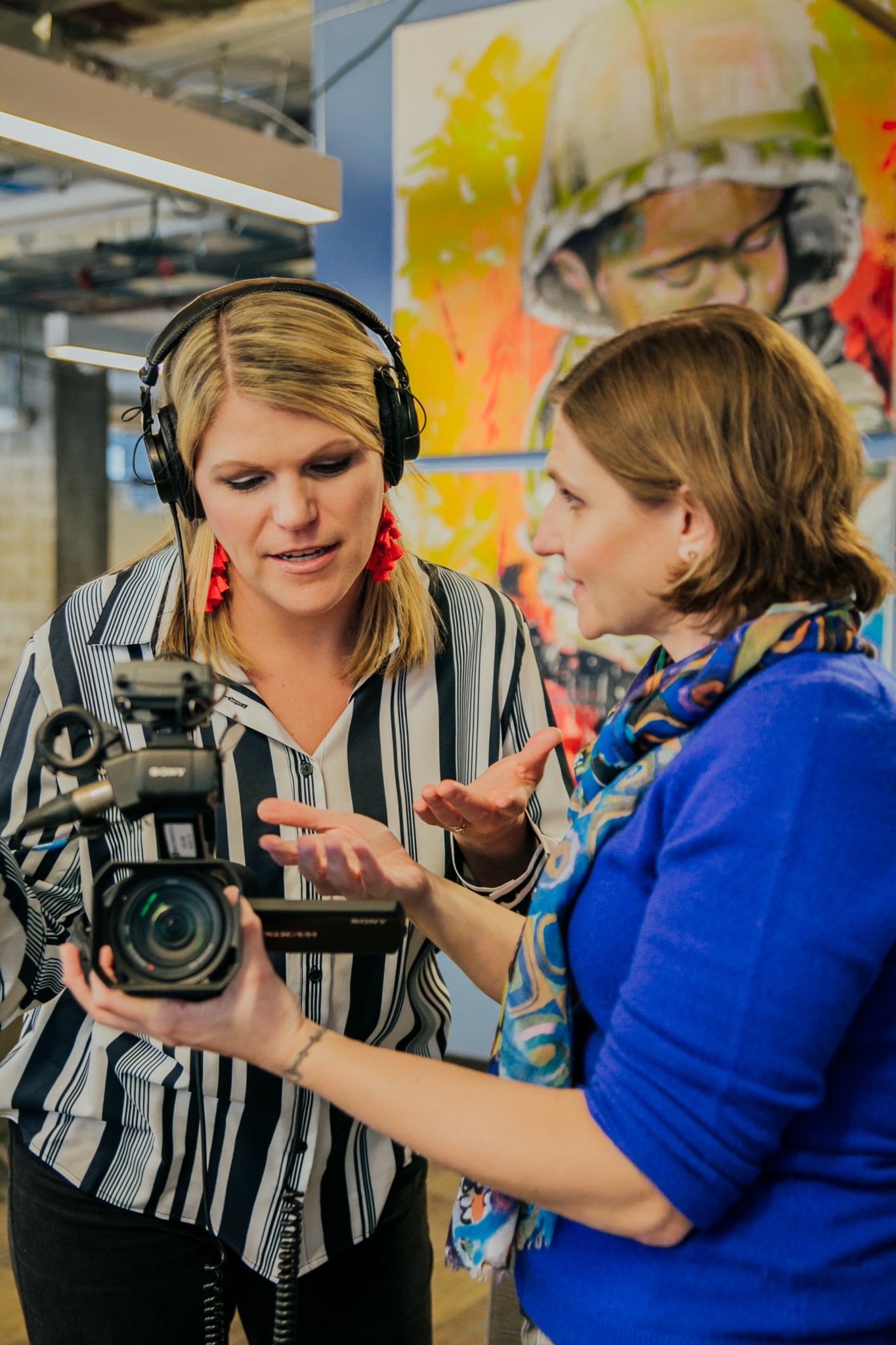 Two women engaged in conversation, one holding a camera, in front of a colorful poster of a person wearing a helmet.