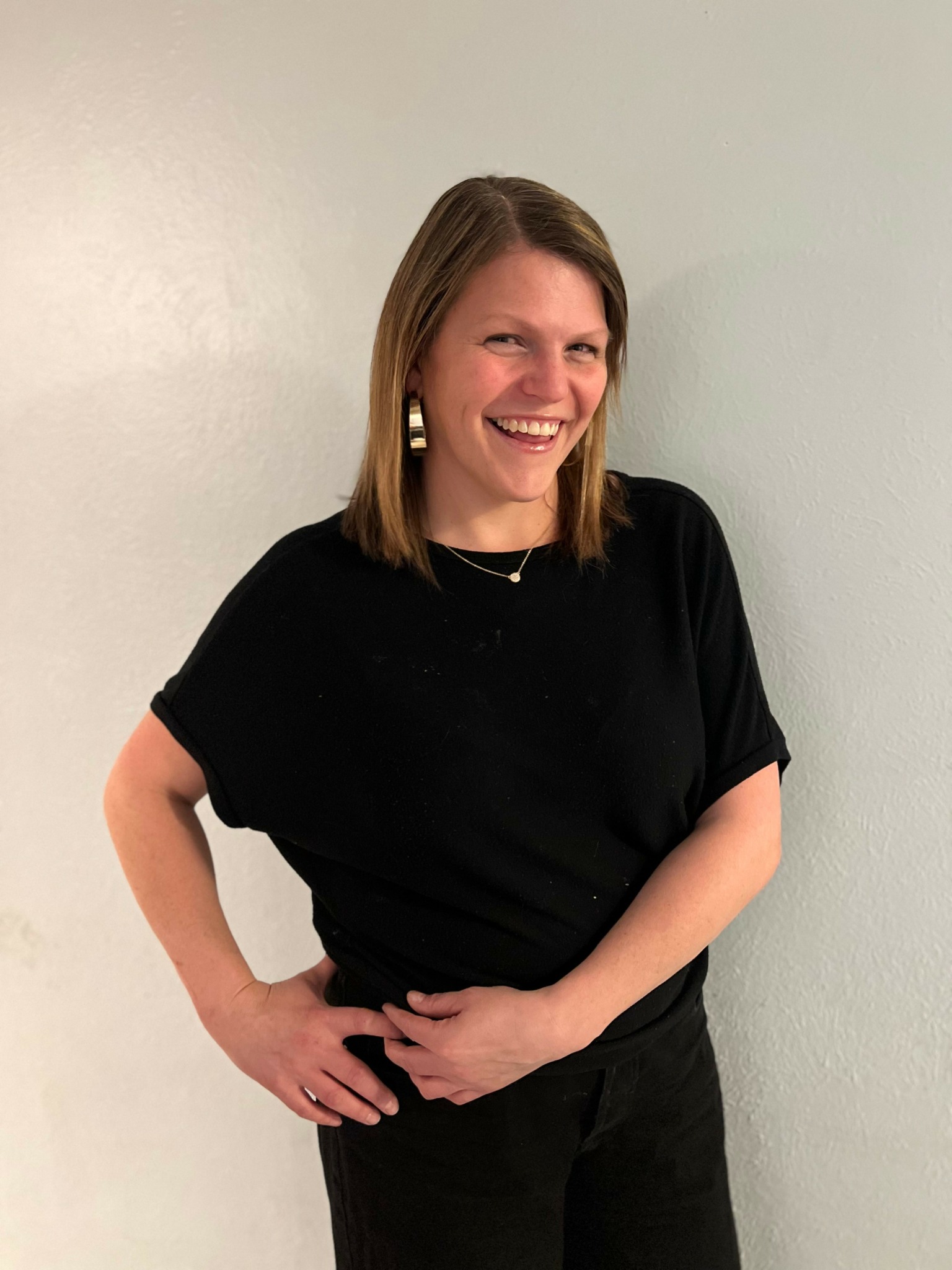 Woman with shoulder-length hair smiling, wearing a black top and black pants, standing against a plain wall.