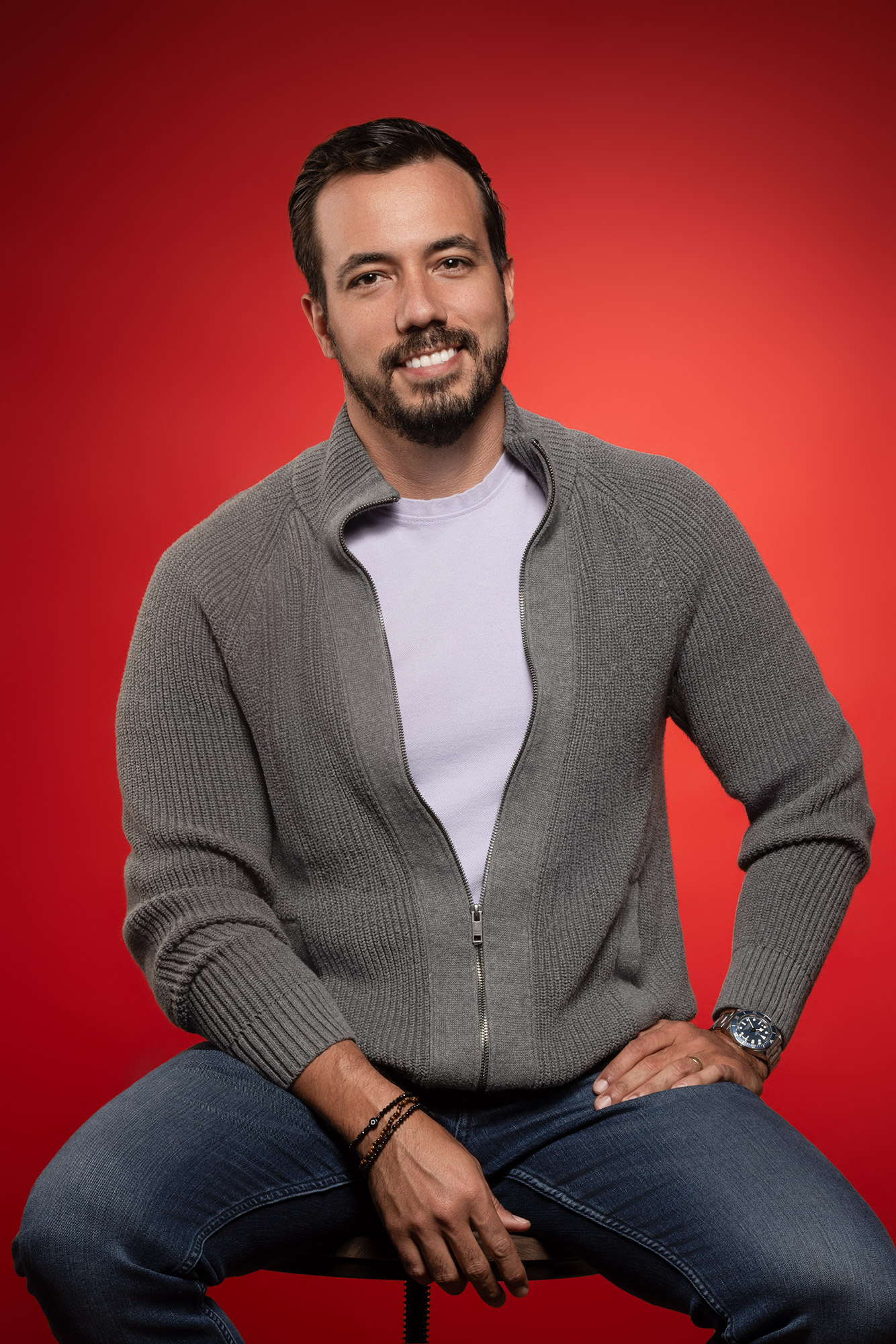 Man with dark hair and beard smiling, wearing a gray zip-up sweater and jeans, sitting against a red background.