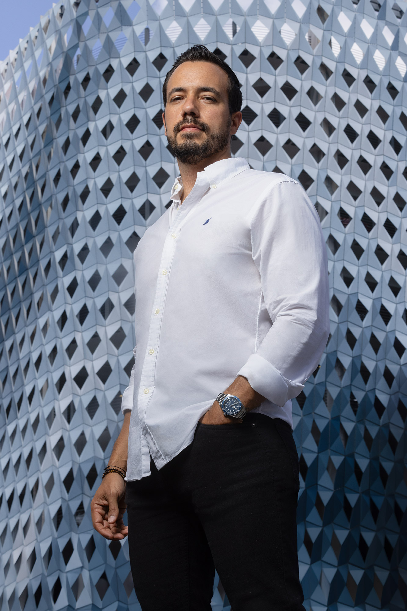 Man with beard in white shirt and black pants stands in front of a geometric patterned wall.