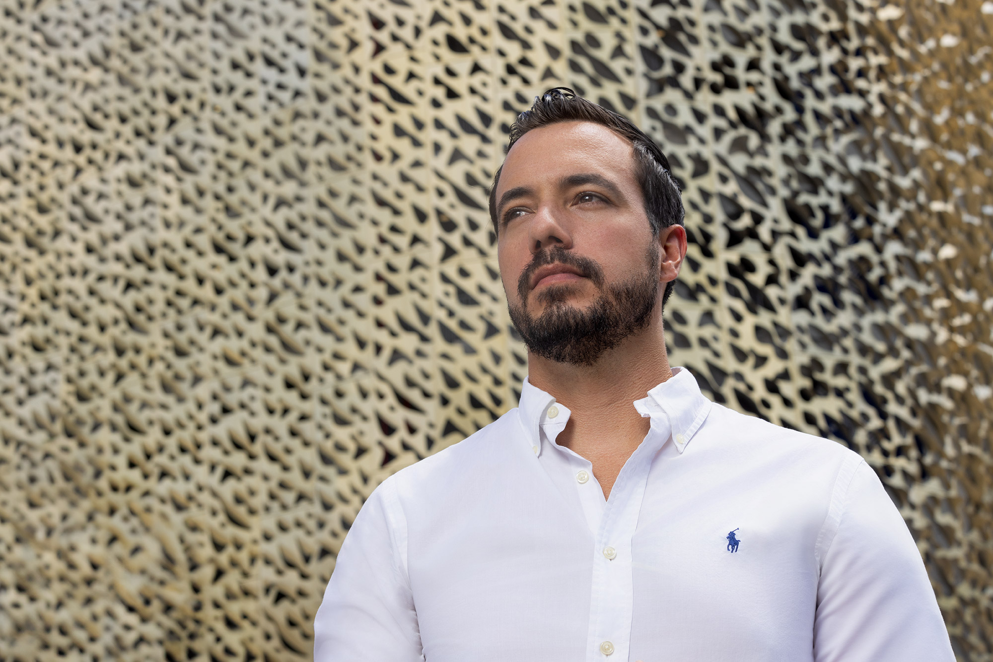 Man with dark hair and beard wearing a white shirt stands outdoors against a textured wall.