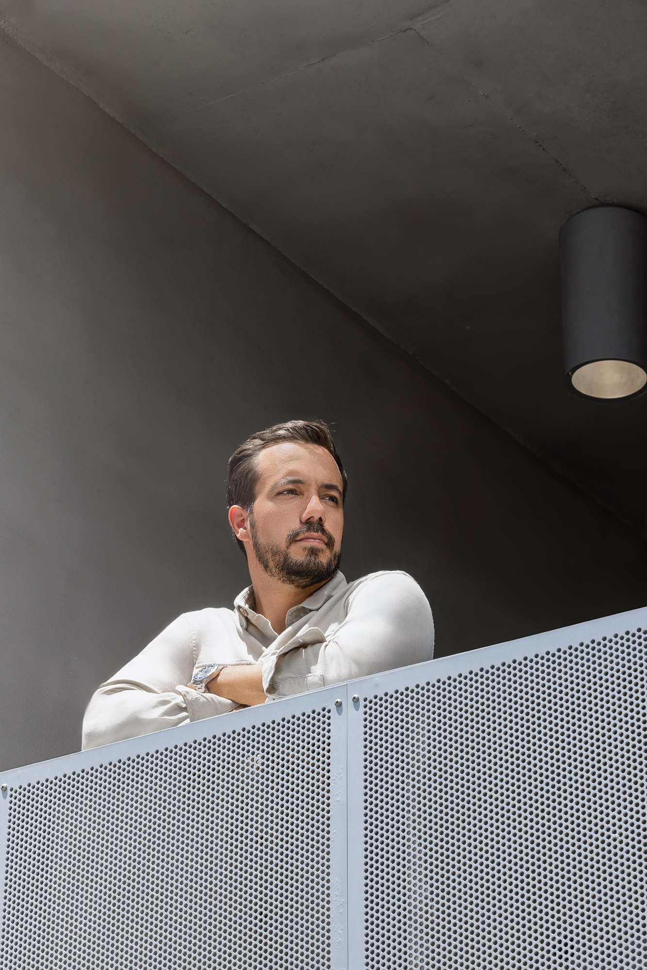 Man with beard and short hair leaning on a perforated metal railing, looking to the side indoors.