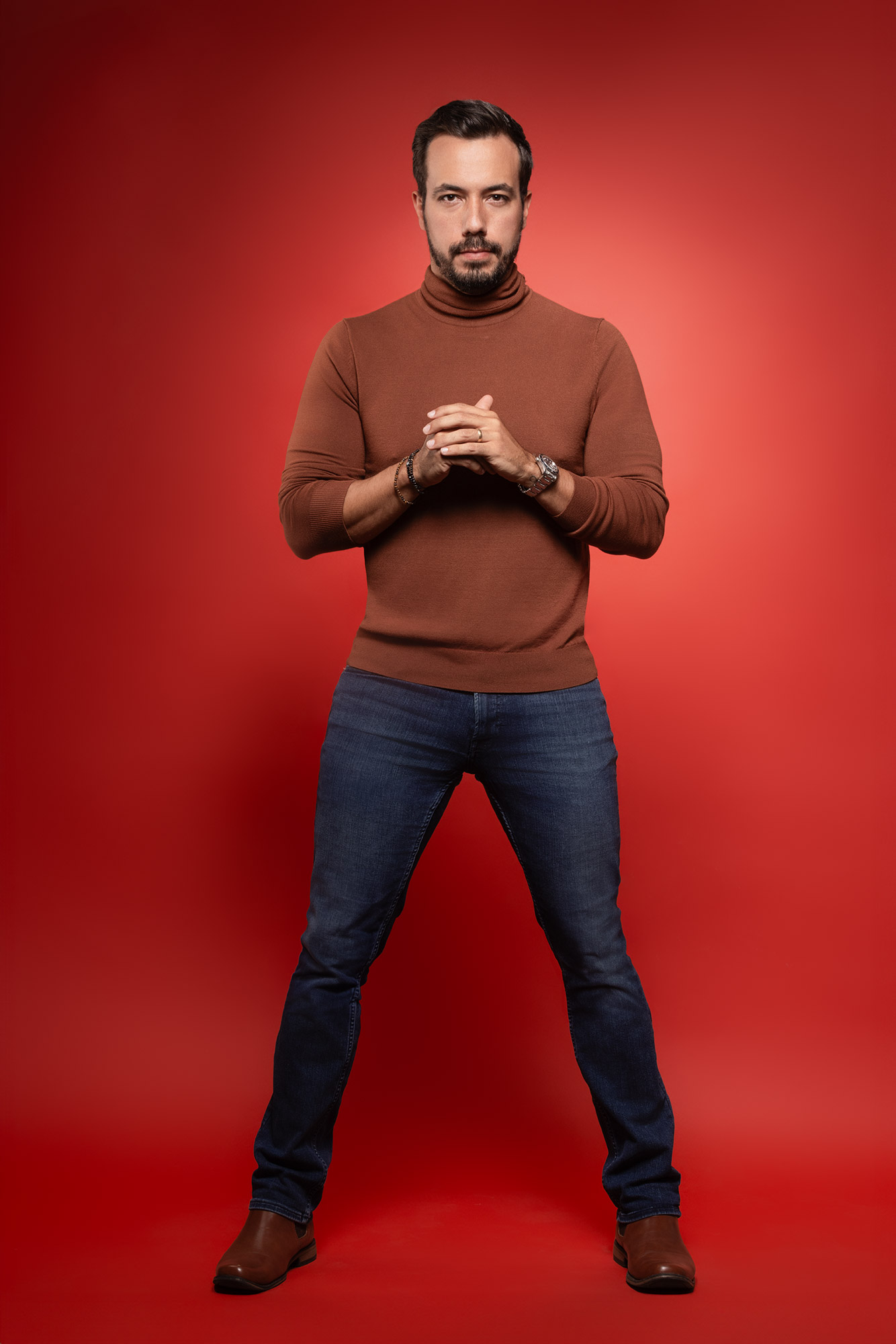 Man with beard and mustache in brown sweater and jeans standing with hands clasped against red background.