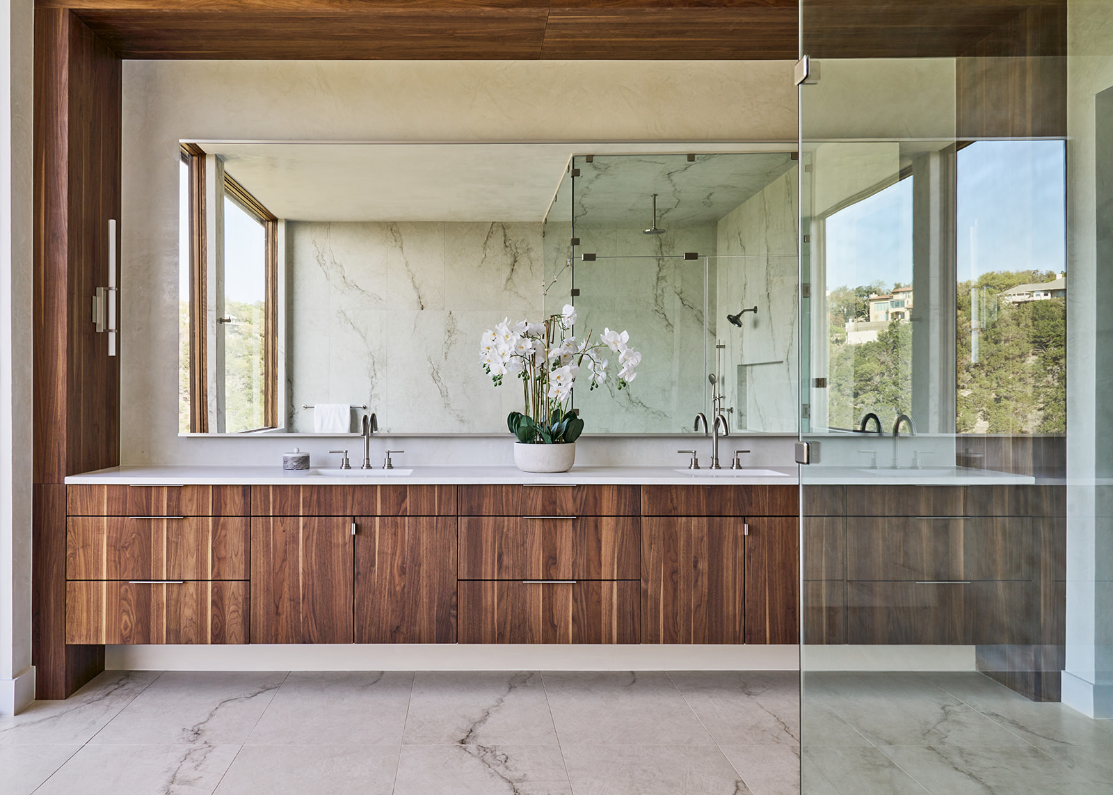 Bathroom vanity with two sinks, wooden cabinet, mirror, and a potted plant, reflected in a large mirror and glass partition.