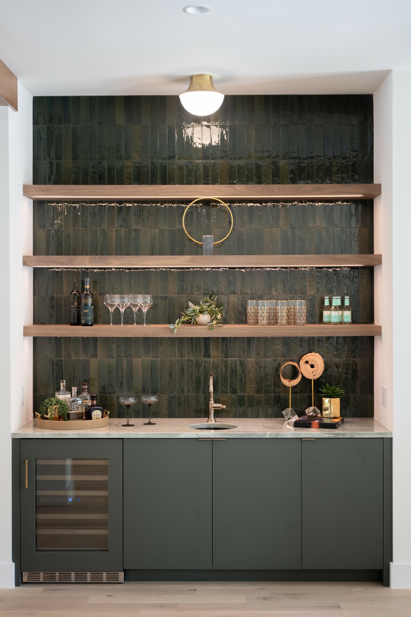 Built-in bar area with dark green tiles, wooden shelves, bottles, plants, and a sink, under a ceiling light.
