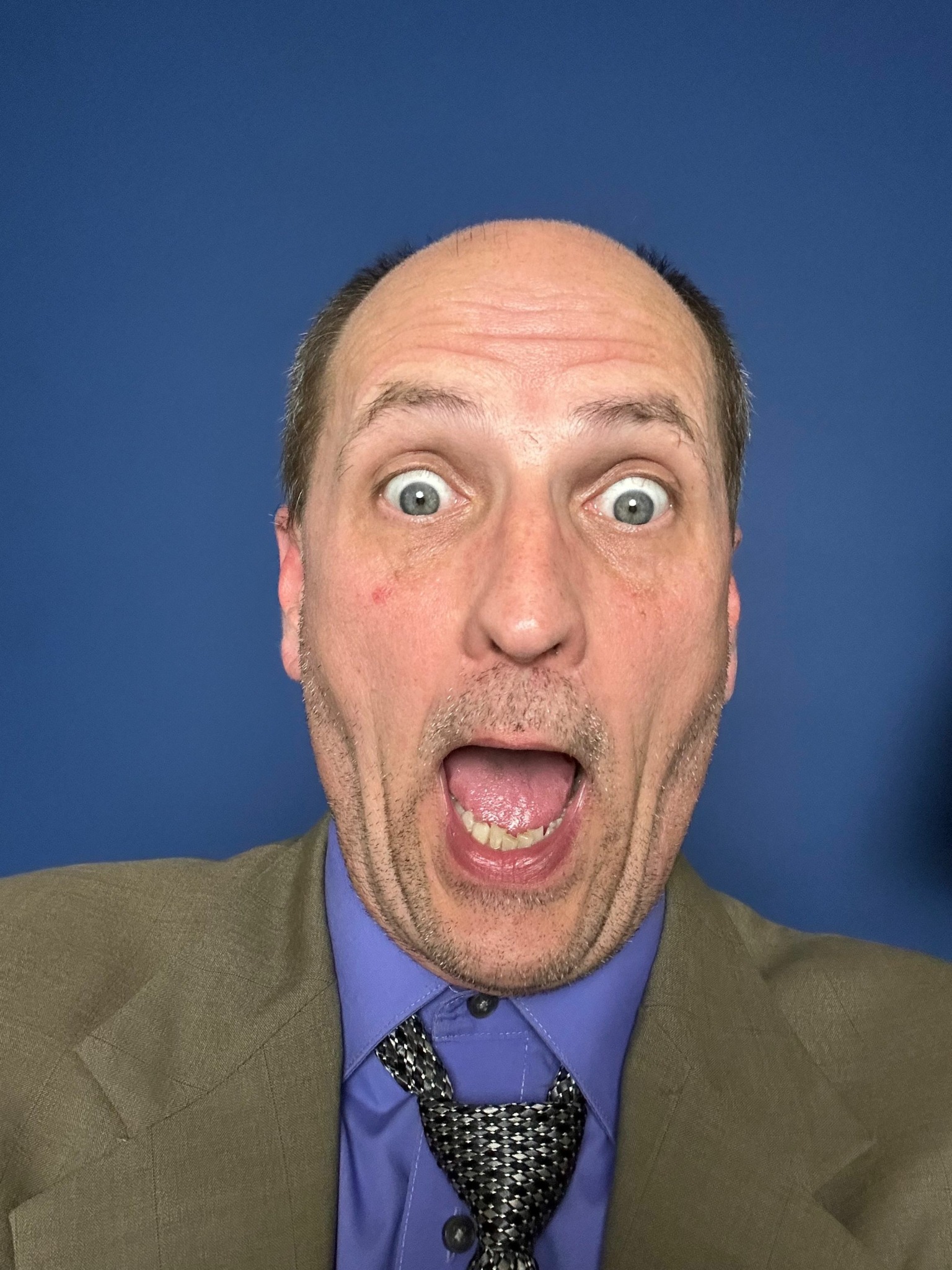 Man with surprised expression, mouth open, eyes wide, wearing a suit and tie, against a blue background.