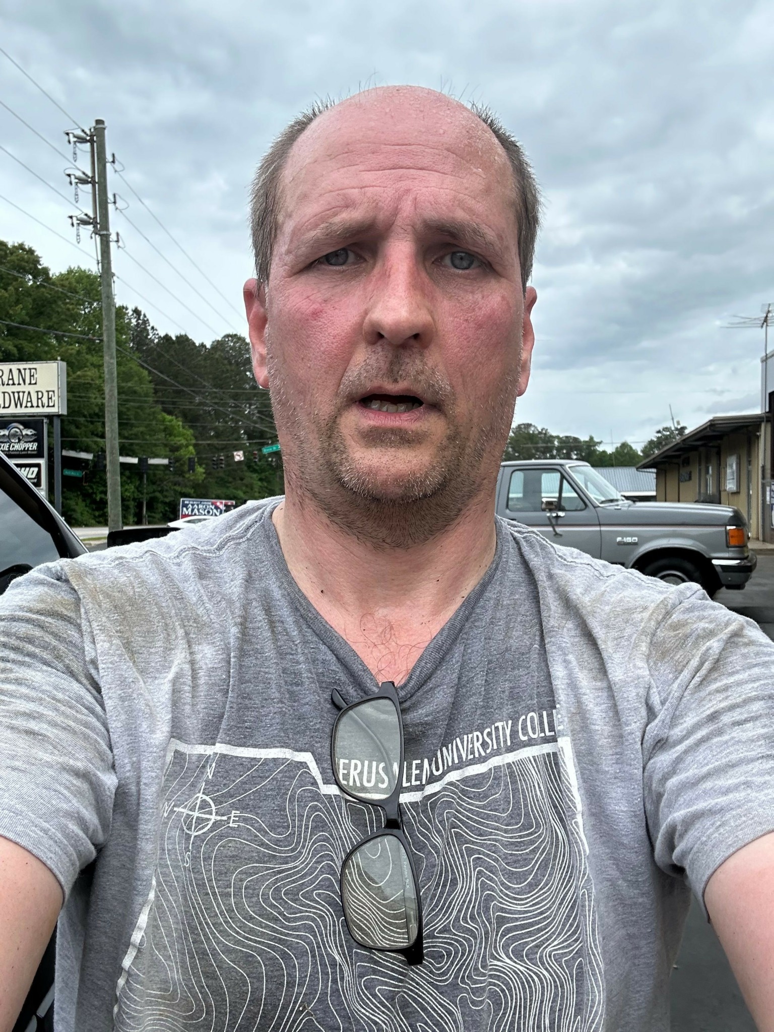 Man with short gray hair wearing a gray shirt outdoors, with cars and trees in background.