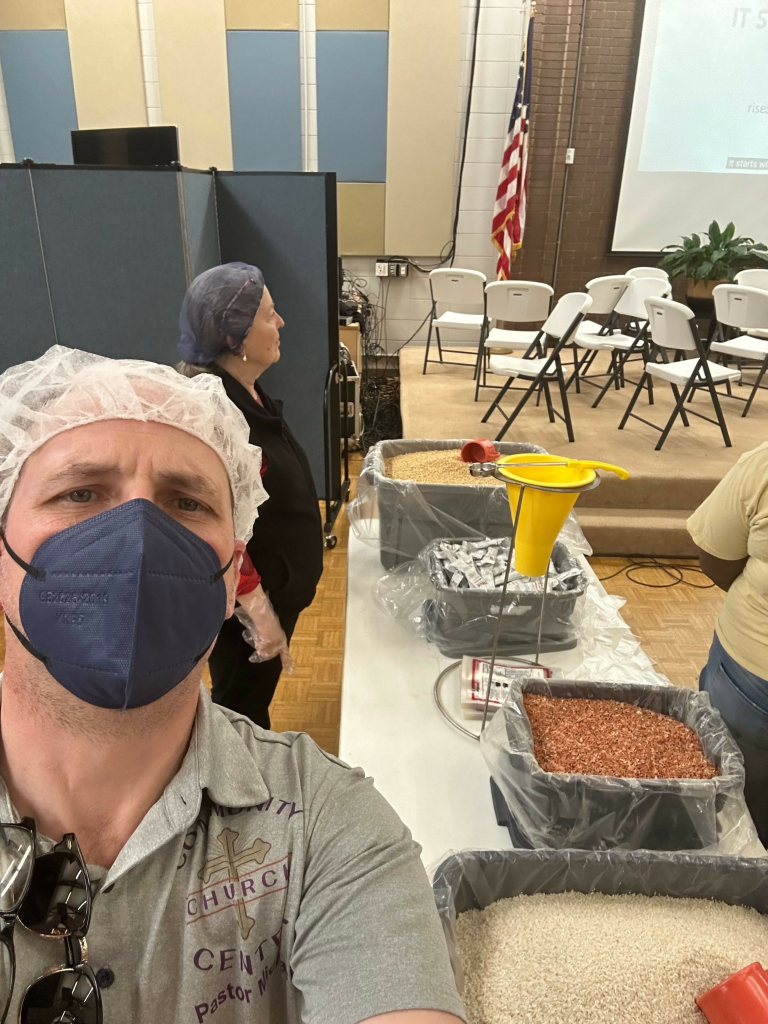 Person wearing a face mask and hairnet, sitting at a table with a yellow funnel and other items, in a room with chairs and an American flag.