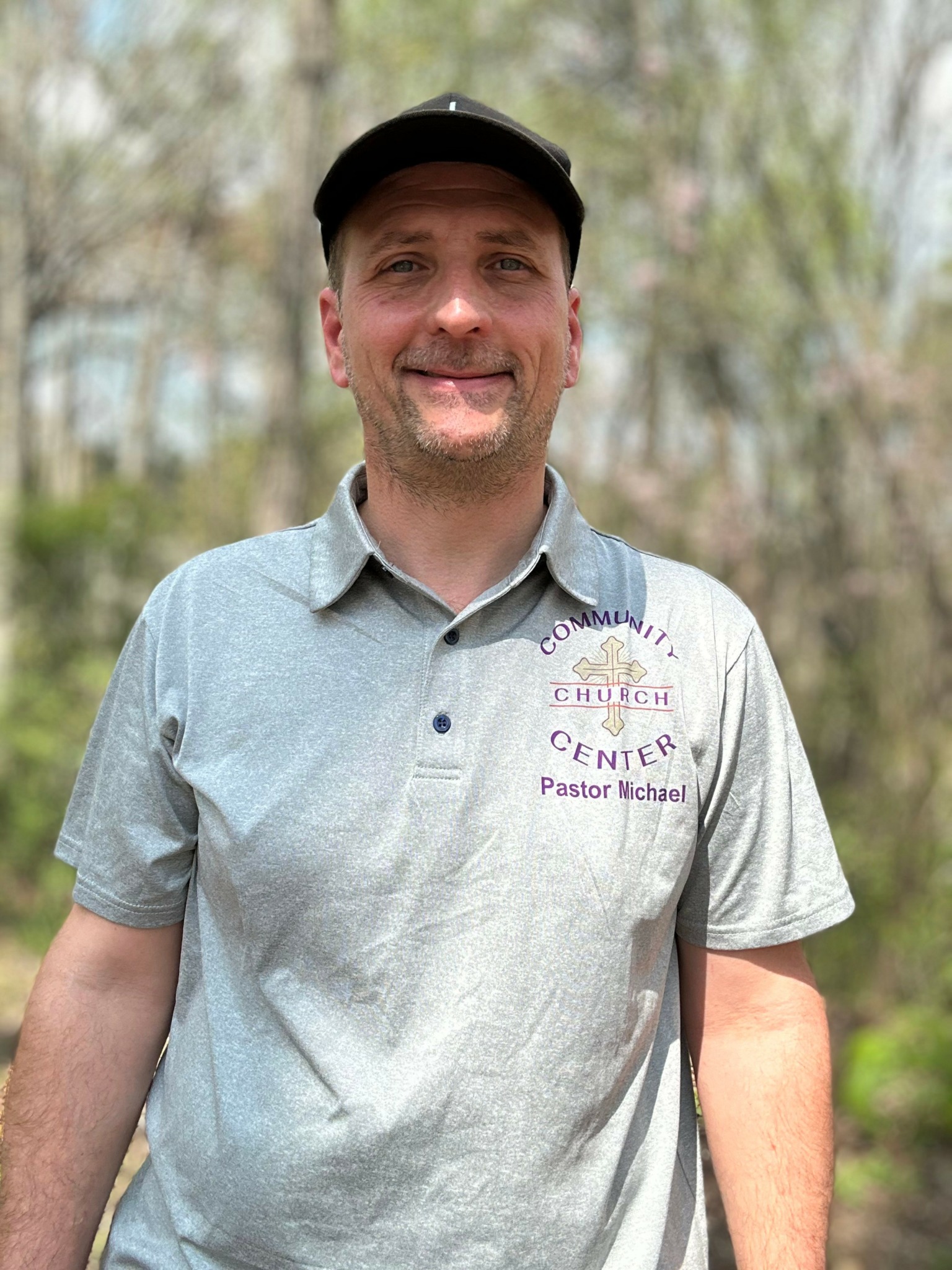 Man smiling outdoors wearing a gray polo shirt with a church logo and black cap.