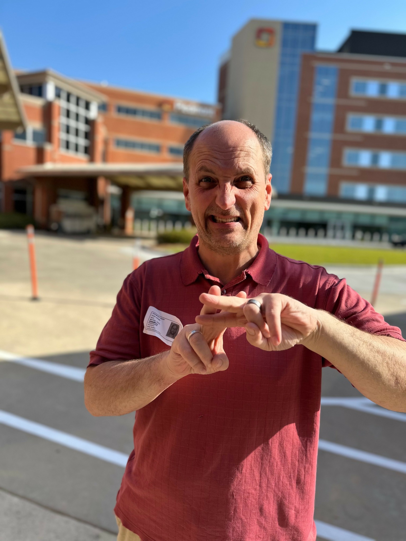 Man with a bald head and a maroon shirt standing outdoors in front of a modern building, smiling.