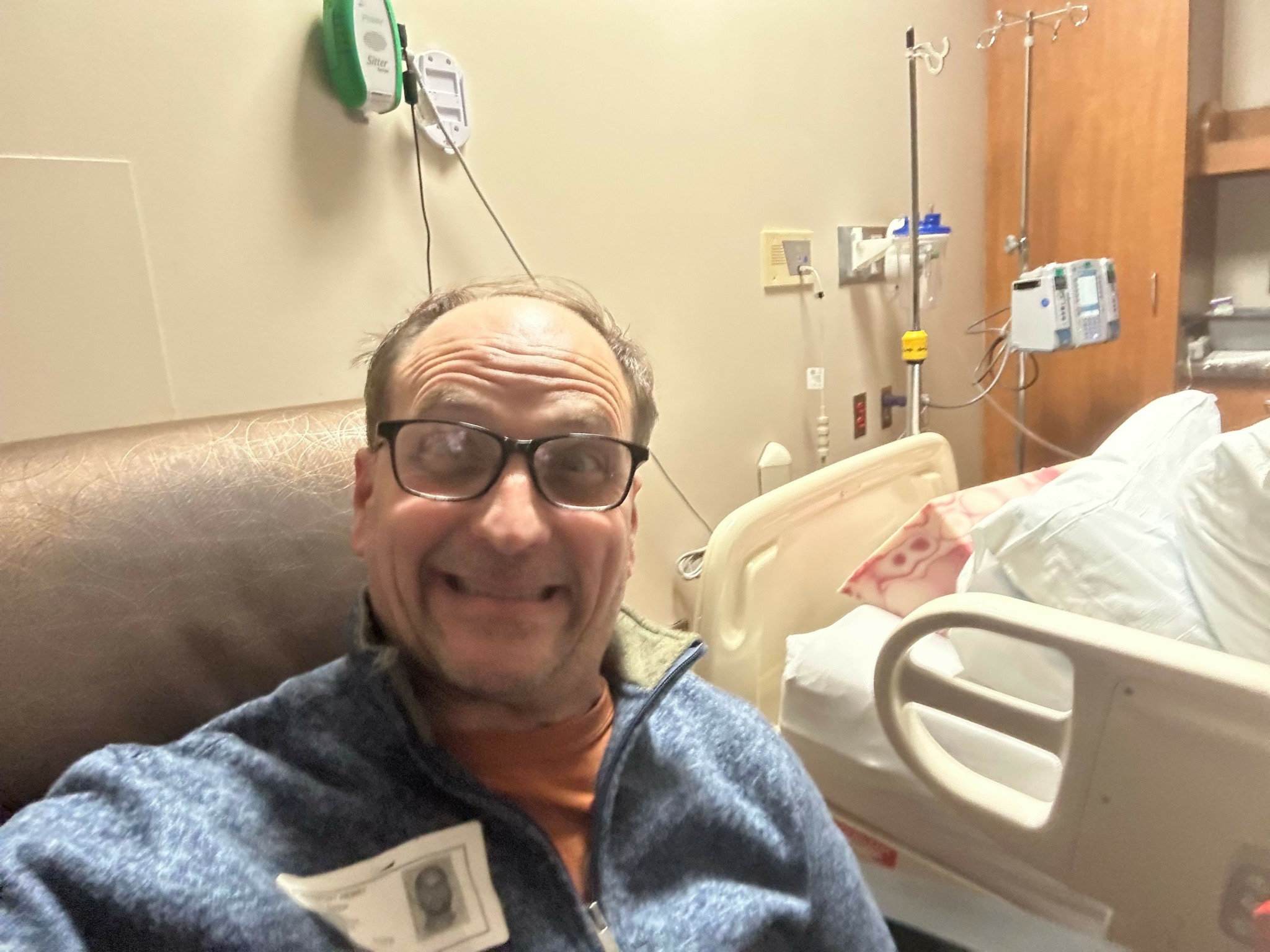 Person with glasses lying on a hospital bed, smiling, with medical equipment nearby.