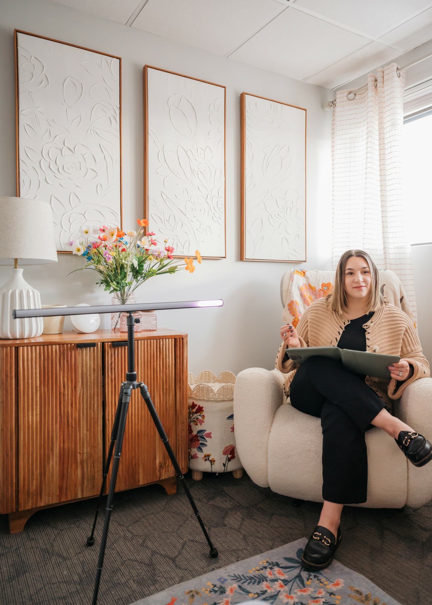 Woman sitting on a white armchair holding a book in a bright room with wall art and a window.