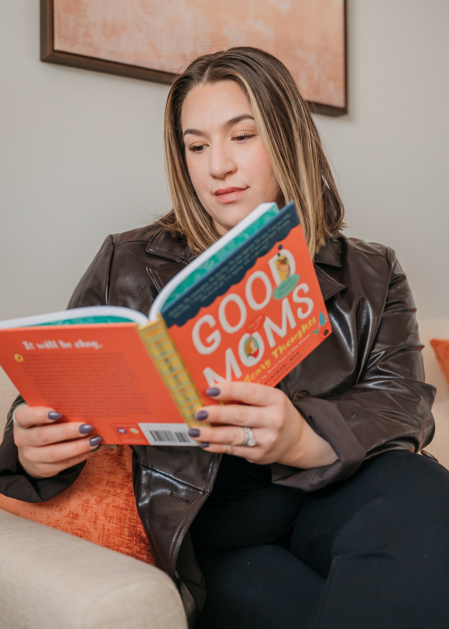 Woman reading a colorful children's book titled 'Good MOMS' while sitting on a couch.