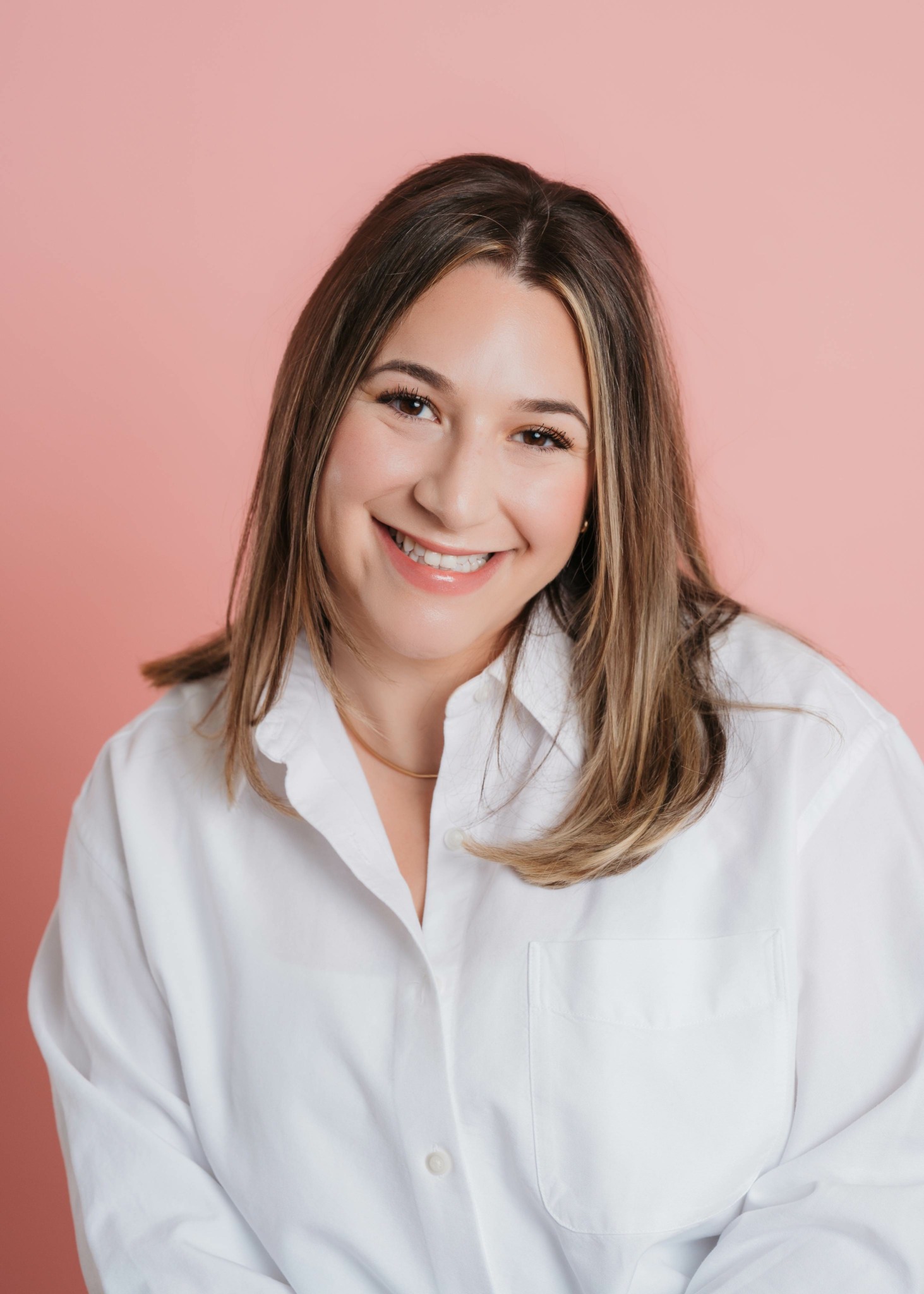 Smiling woman with shoulder-length hair wearing a white shirt against pink background.