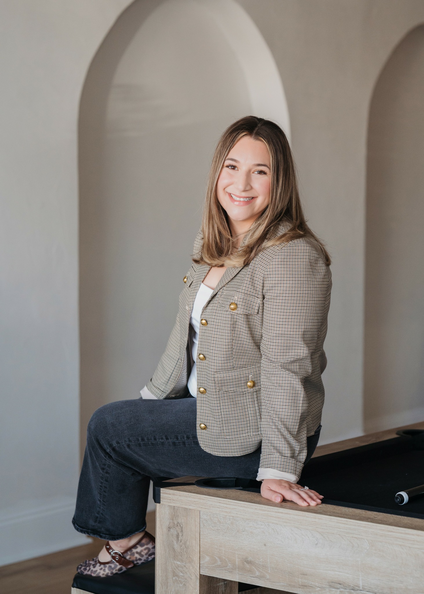 Young woman with shoulder-length hair smiling, sitting on a wooden surface, wearing a checkered blazer and dark jeans.