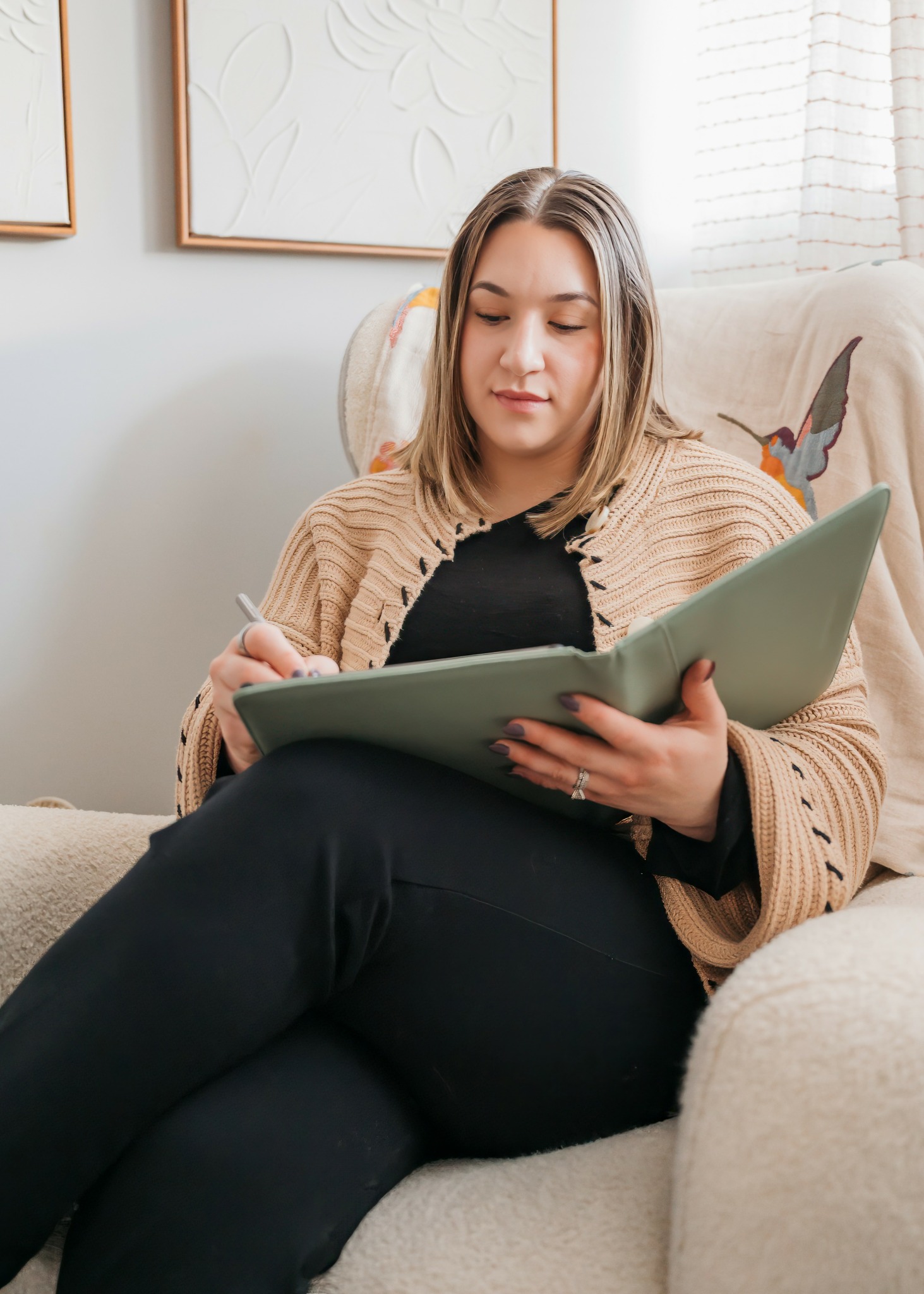 Woman sitting on sofa, writing in notebook, in a bright room with framed art and butterfly pillow.