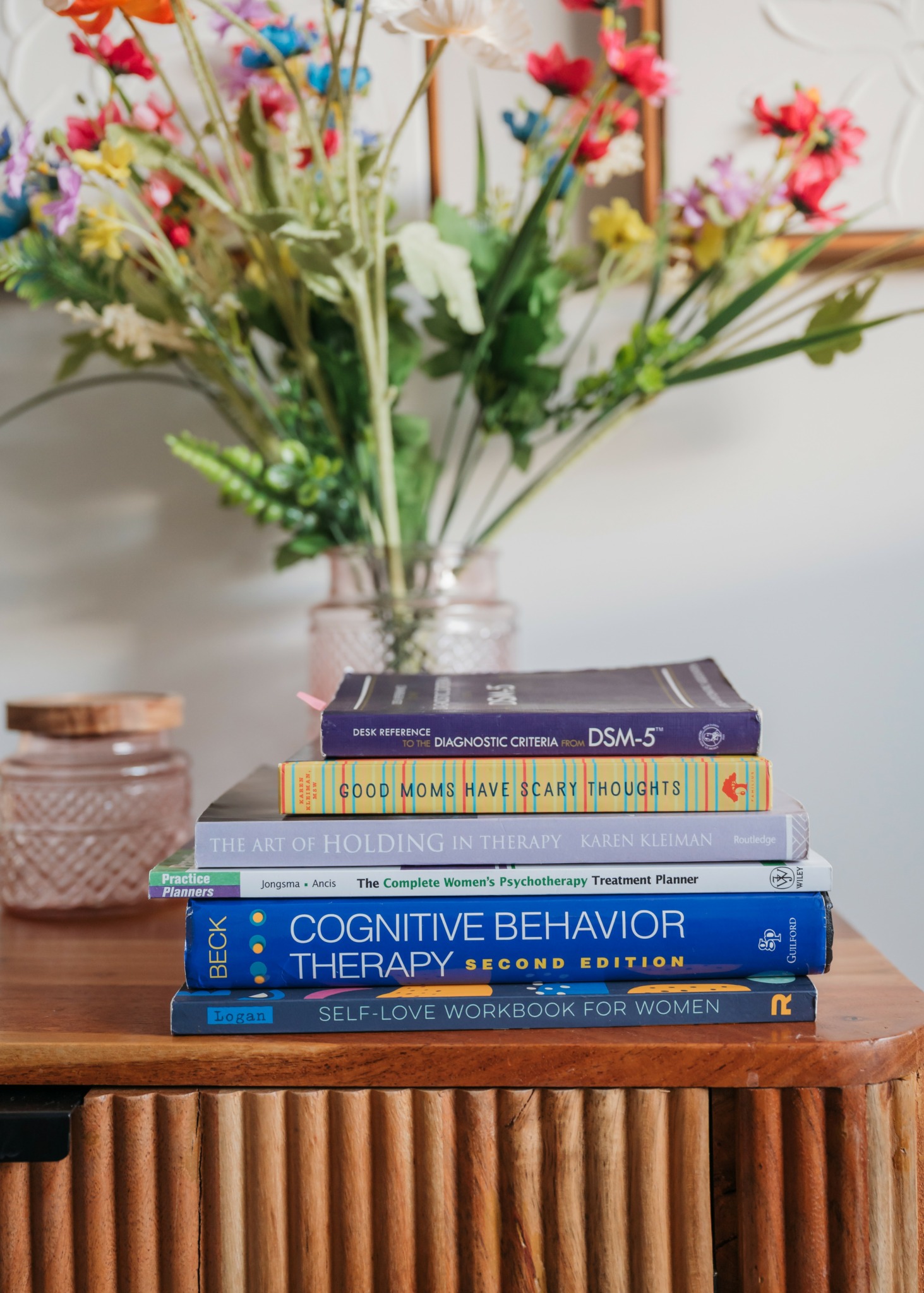 Stack of books on a wooden table with a vase of colorful flowers behind them.