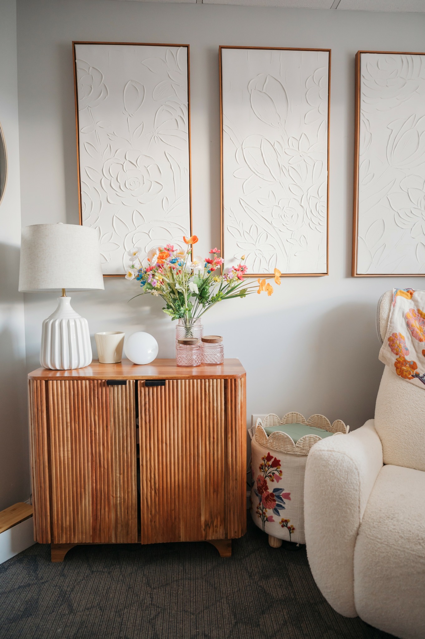 Wooden cabinet with a lamp, flowers in a vase, and decorative objects, next to a white armchair with floral cushion.
