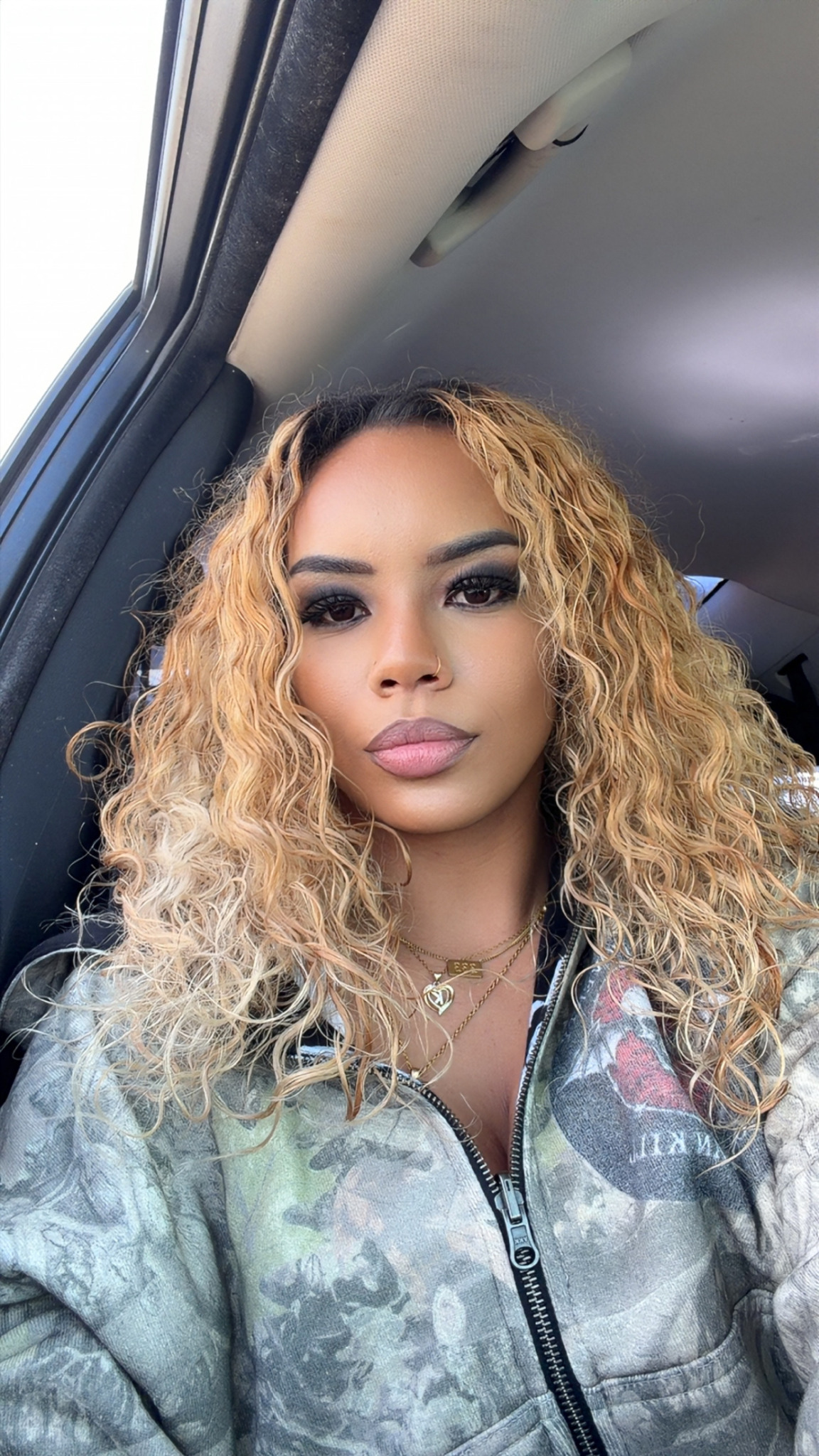 Young woman with curly blonde hair and makeup, sitting inside a car, wearing a camouflage jacket.