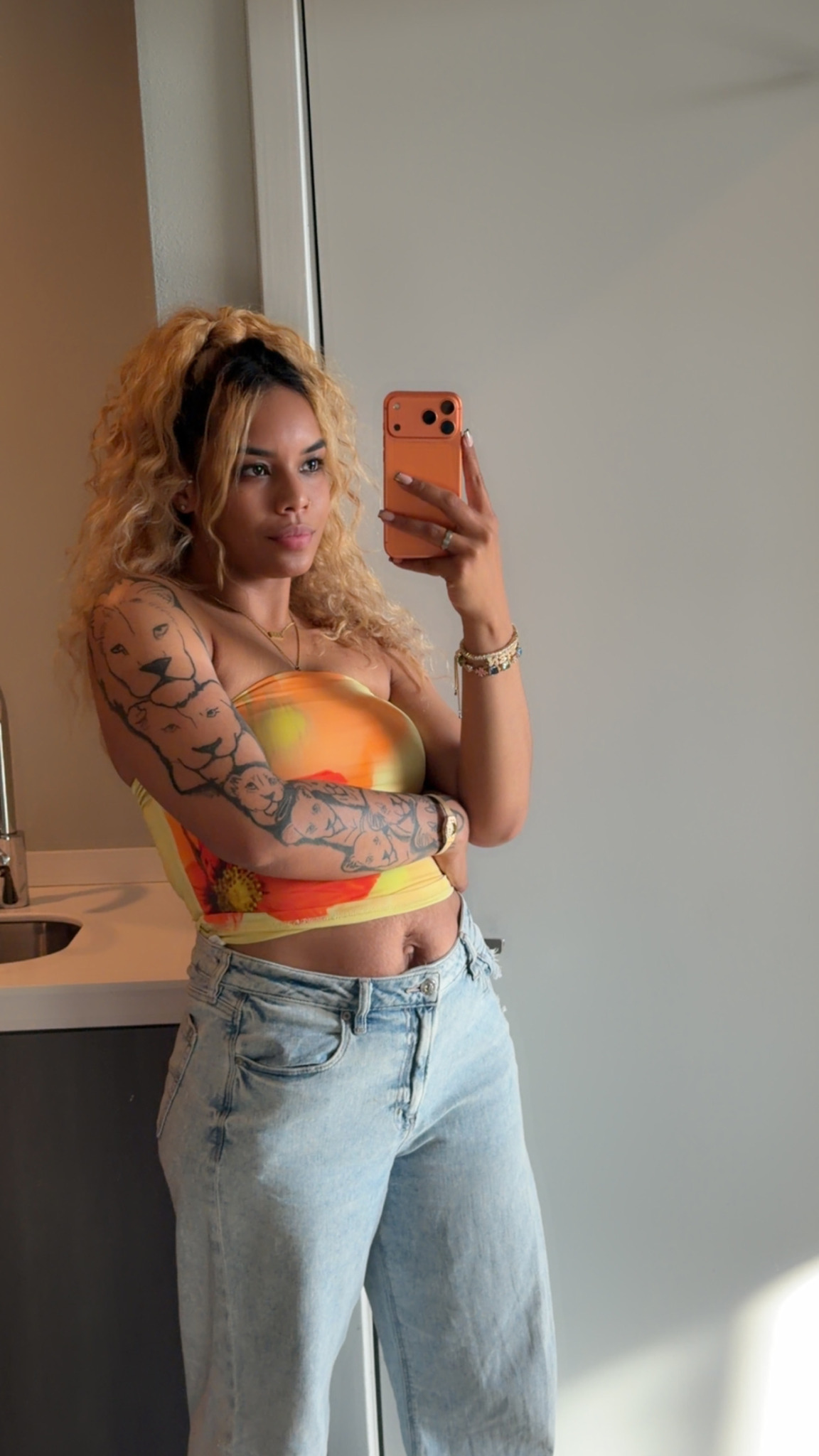 Young woman with curly hair taking a mirror selfie, wearing a colorful crop top and light jeans.