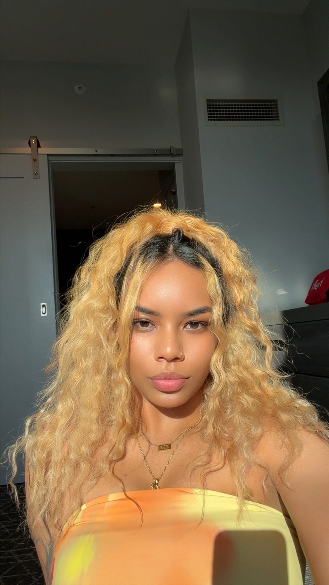 Young woman with long, curly blonde hair taking a selfie indoors, looking at the camera with a neutral expression.