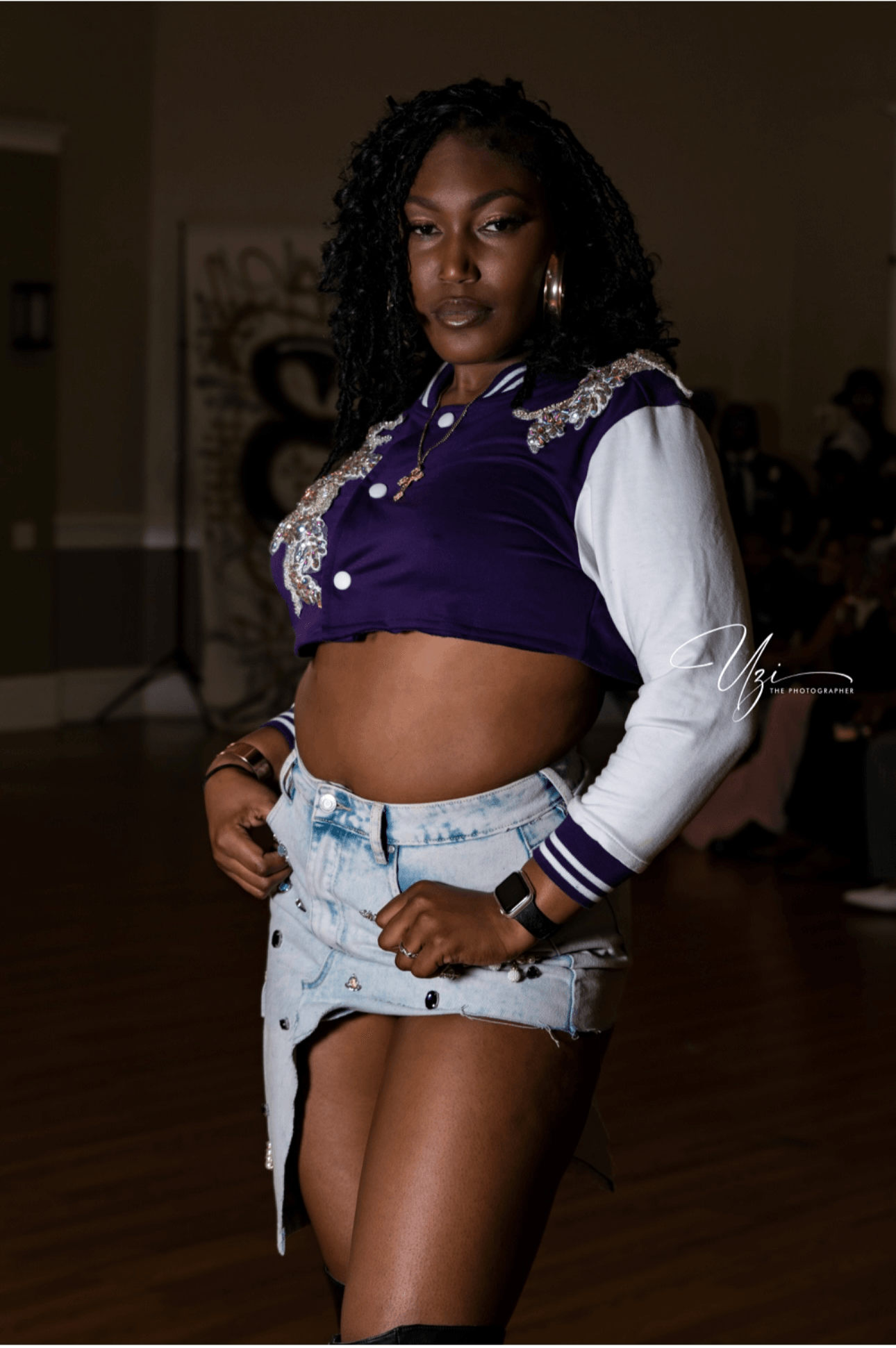 Young woman with curly hair wearing a purple and white jacket and denim skirt, standing indoors.