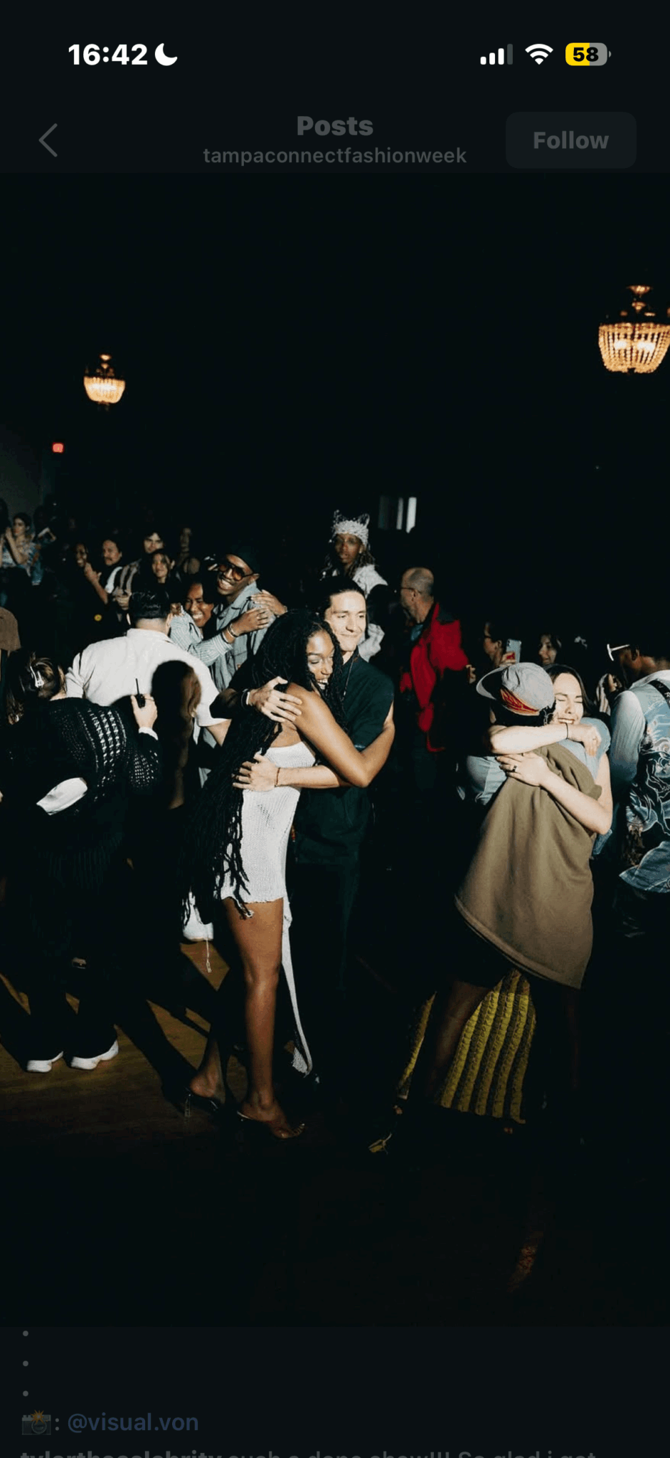 People dancing and socializing in a dark room with chandeliers, some embracing and holding drinks.