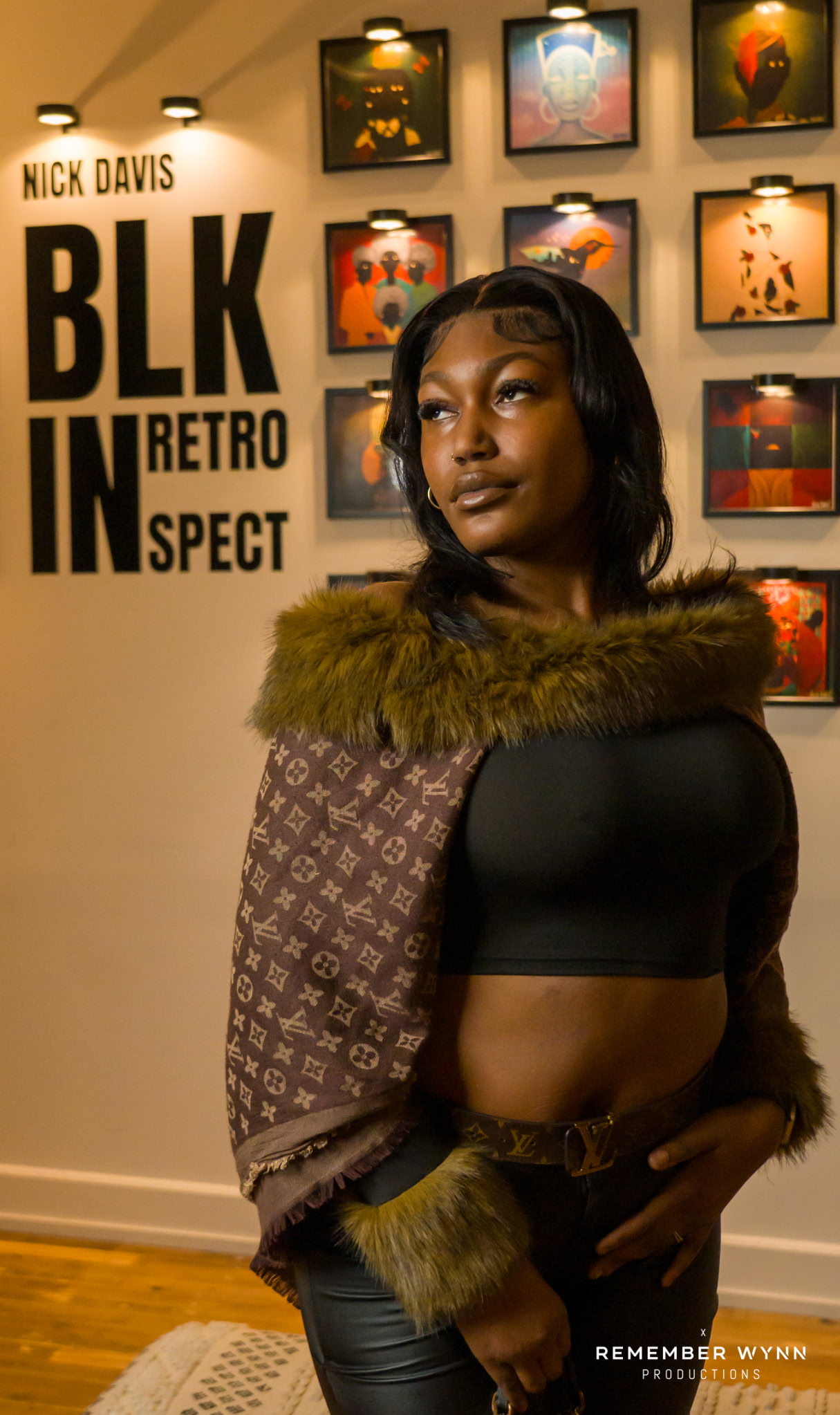 Woman with dark hair wearing a black crop top and fur-lined jacket, standing in front of art display wall.