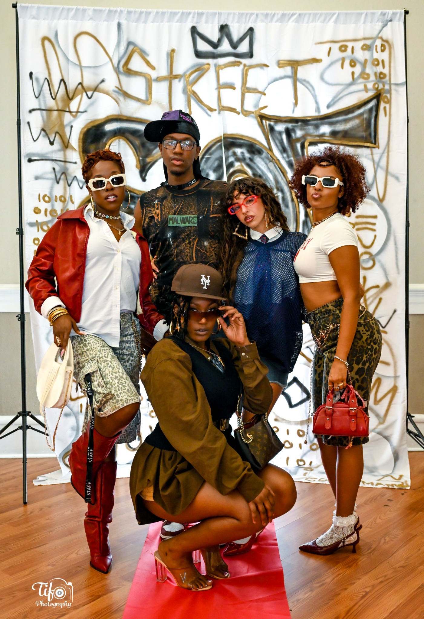 Five people pose in front of graffiti backdrop, wearing stylish clothing and sunglasses, with one person crouching in front.