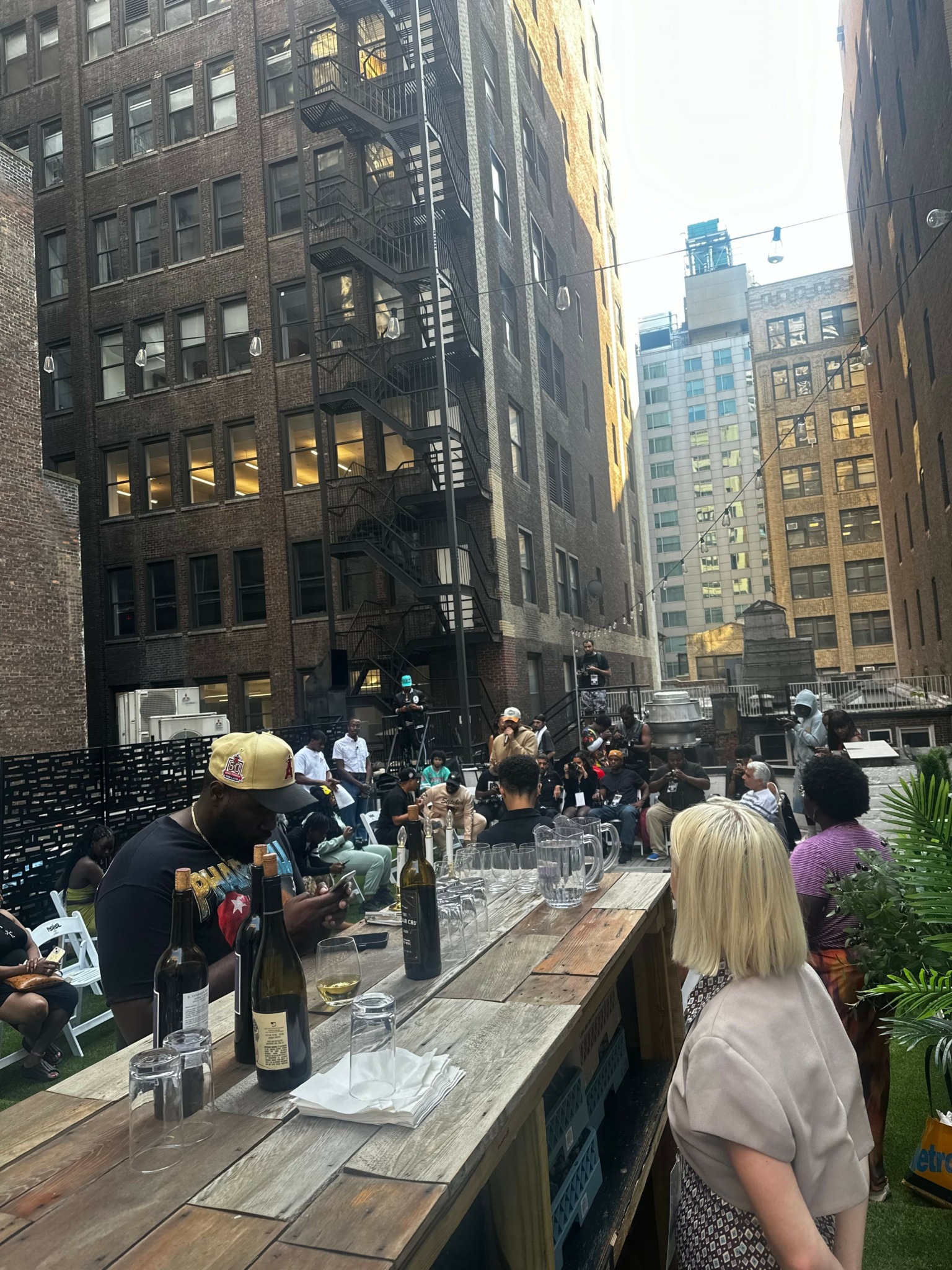 Street scene with people, buildings, and outdoor seating area, viewed from a sidewalk with a wooden railing.