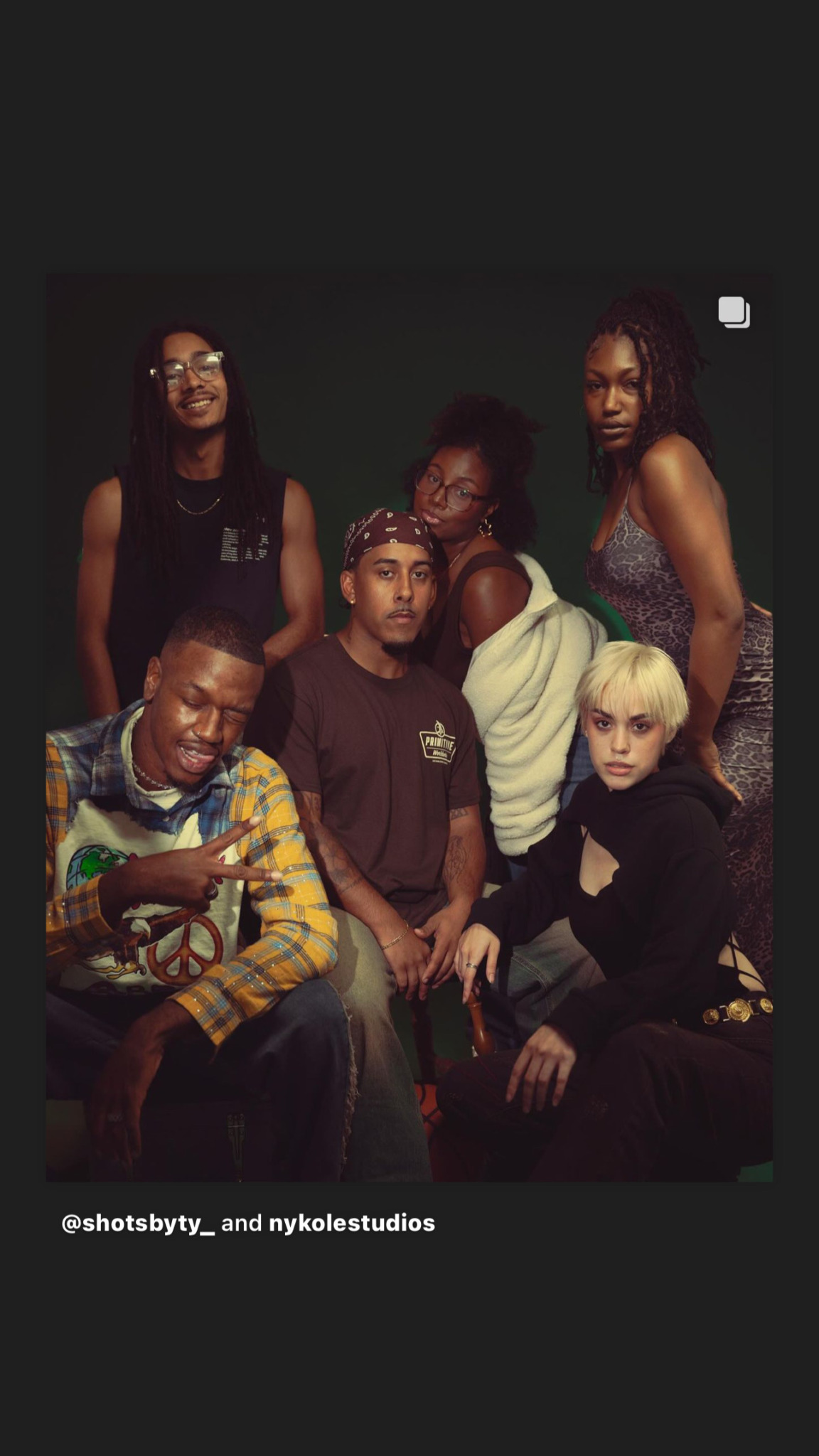 Six people pose together against a dark background, some sitting and some standing, with varied clothing styles.