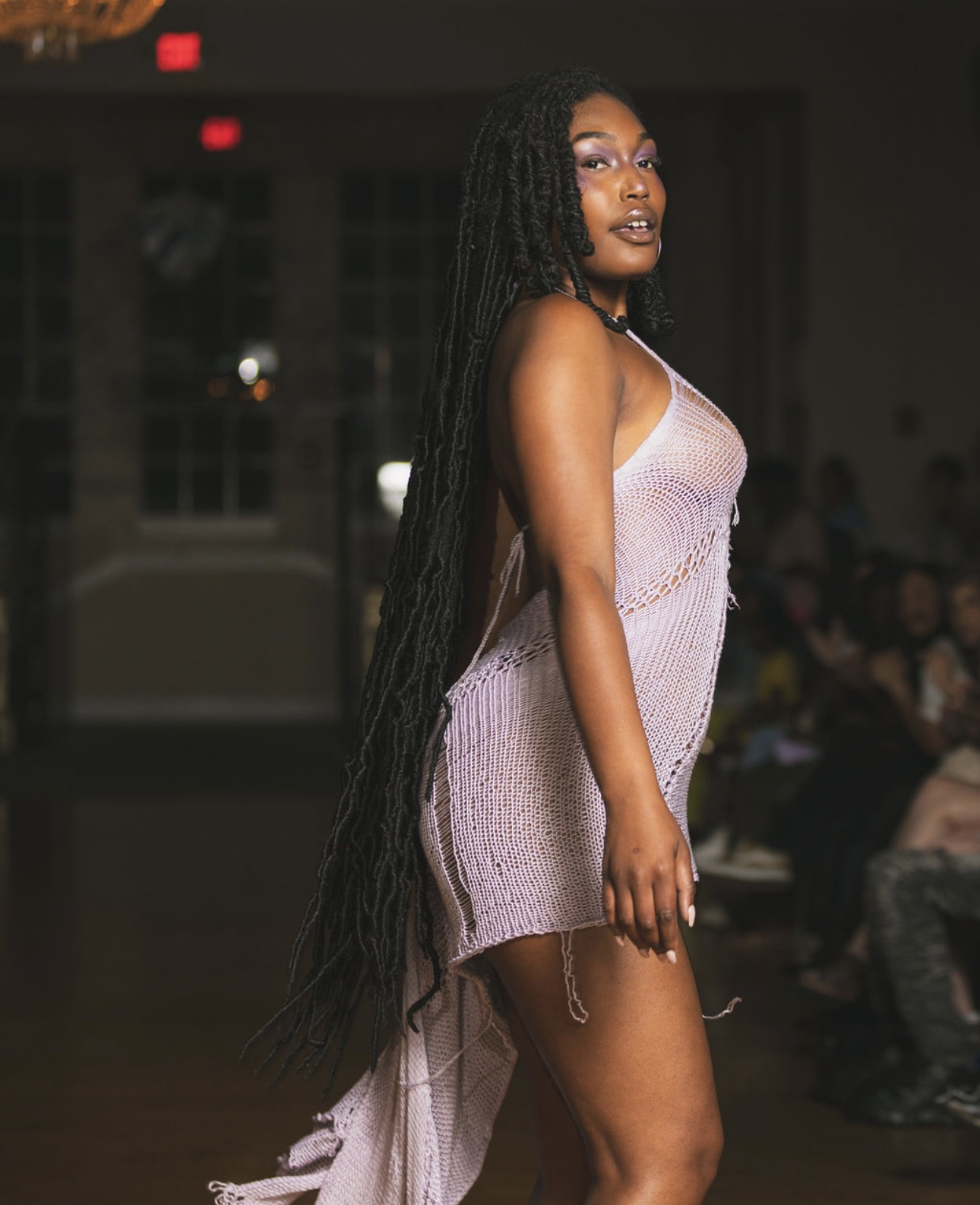Woman with long braids in a sleeveless dress standing indoors, audience in background.