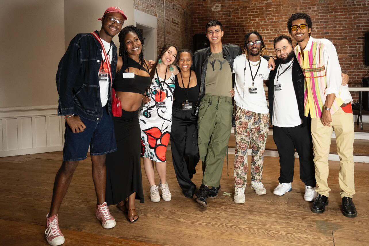 Group of seven people standing together indoors, smiling, with brick wall background and wooden floor.