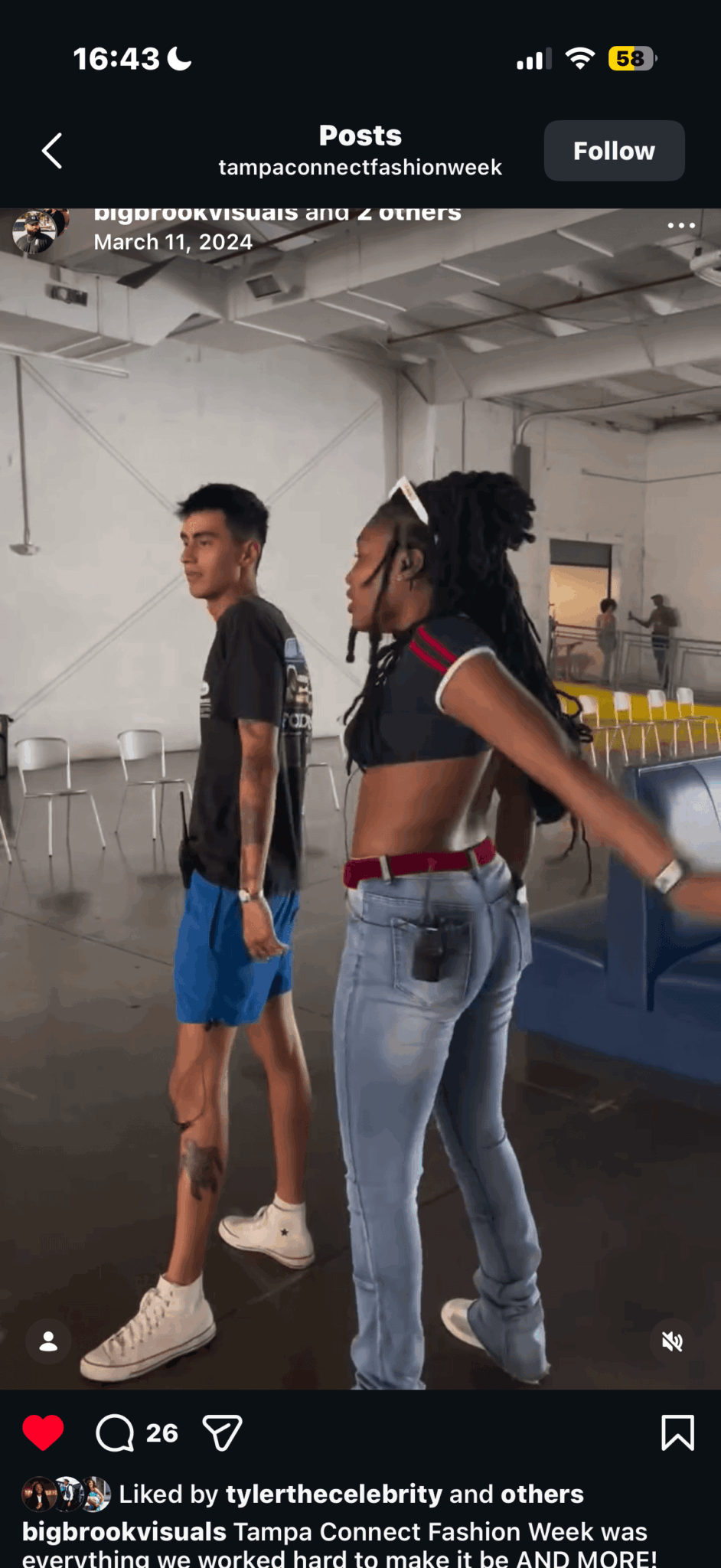 Two young people standing in a room with chairs and tables, one with hands on hips, the other looking away.