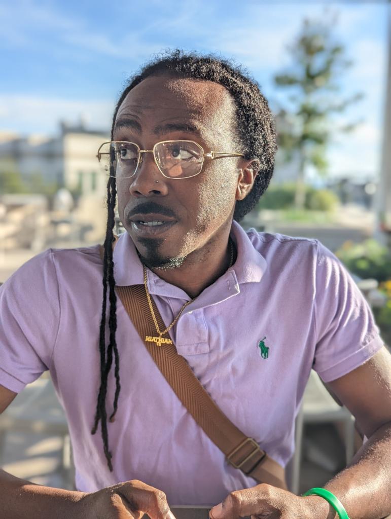Man with glasses and long dreadlocks wearing a purple polo shirt outdoors.
