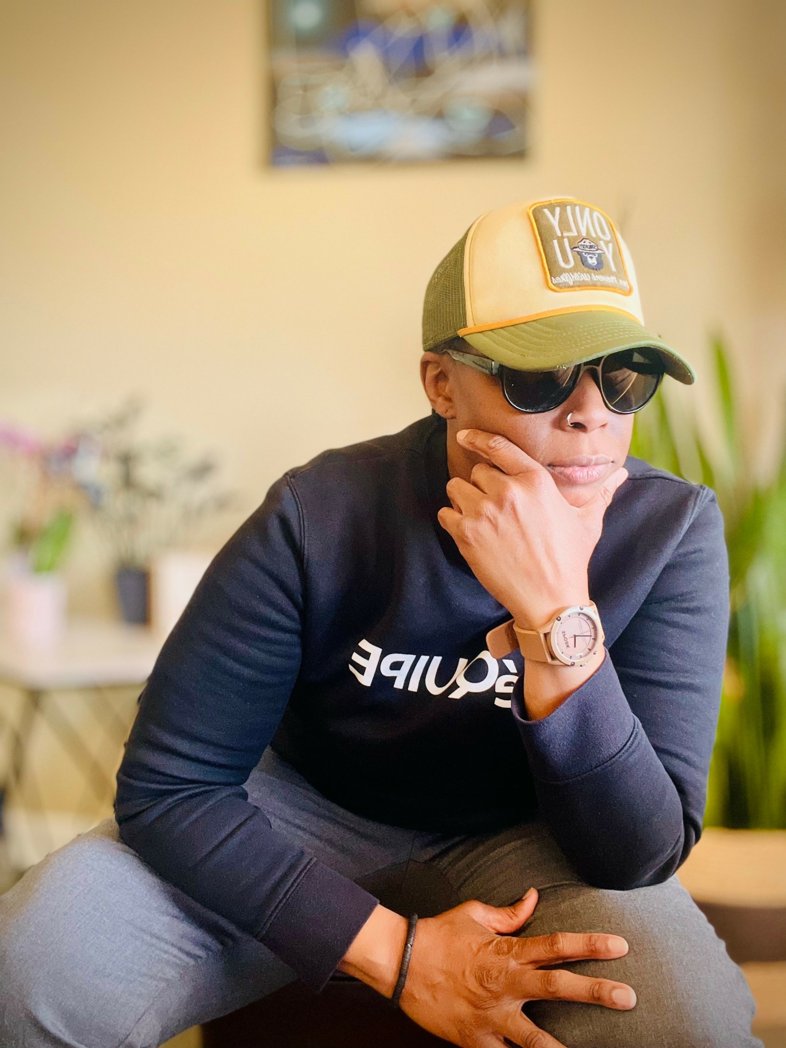 Person wearing sunglasses, cap, and dark sweatshirt, sitting with hand on chin indoors.