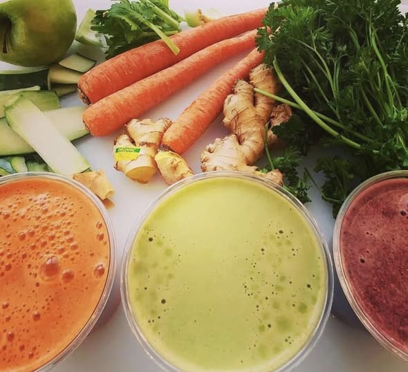 Assorted fresh vegetables and three smoothies in glasses, including carrots, ginger, leafy greens, and other vegetables.